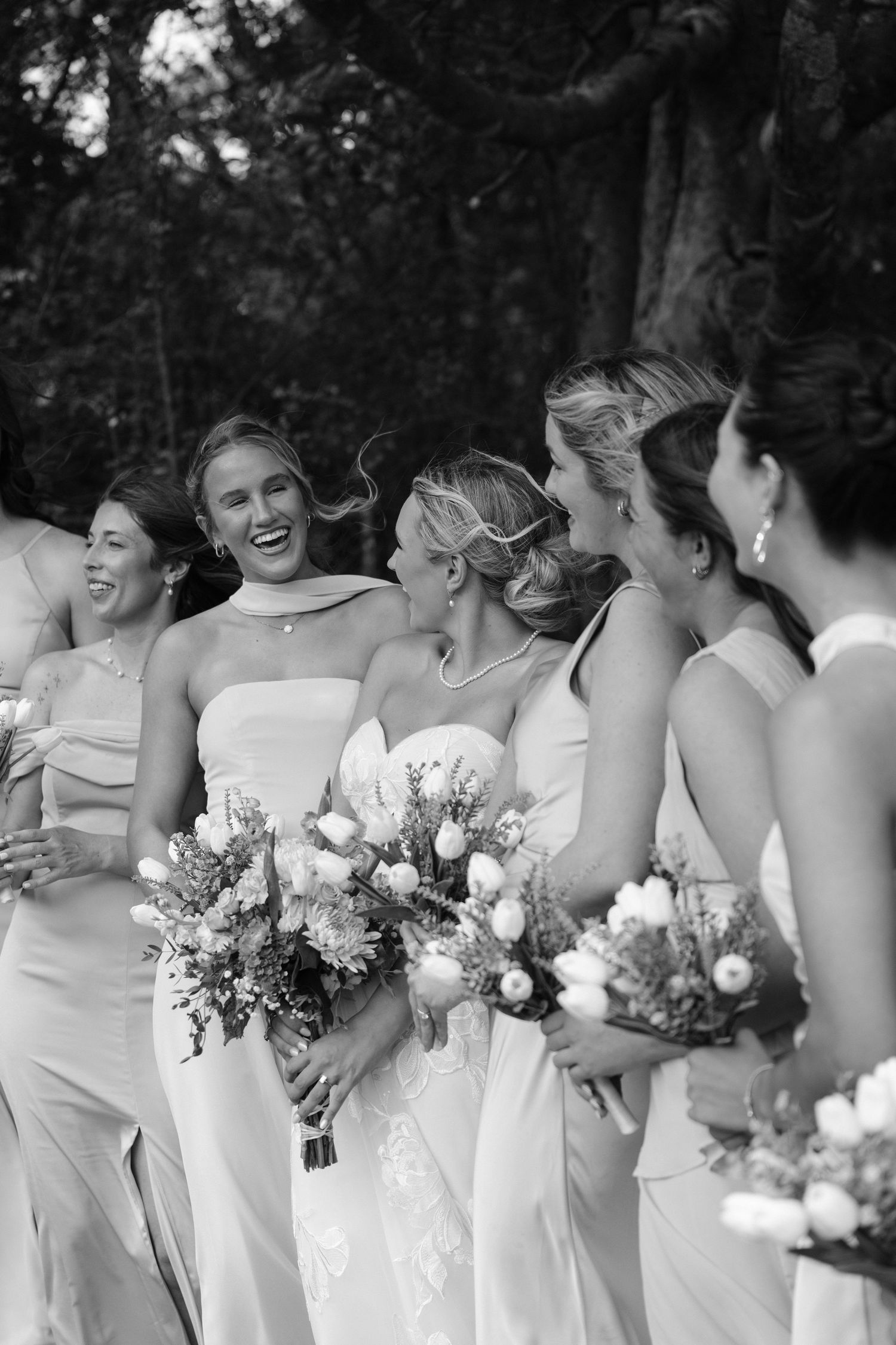 Bride and bridesmaids in white dresses laughing together outdoors holding floral bouquets.