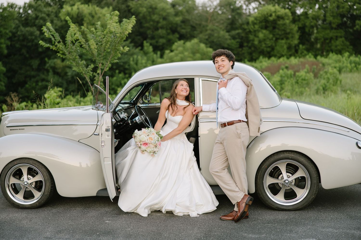 Bride and groom posing with a vintage cream classic car on their wedding day, surrounded by lush greenery.