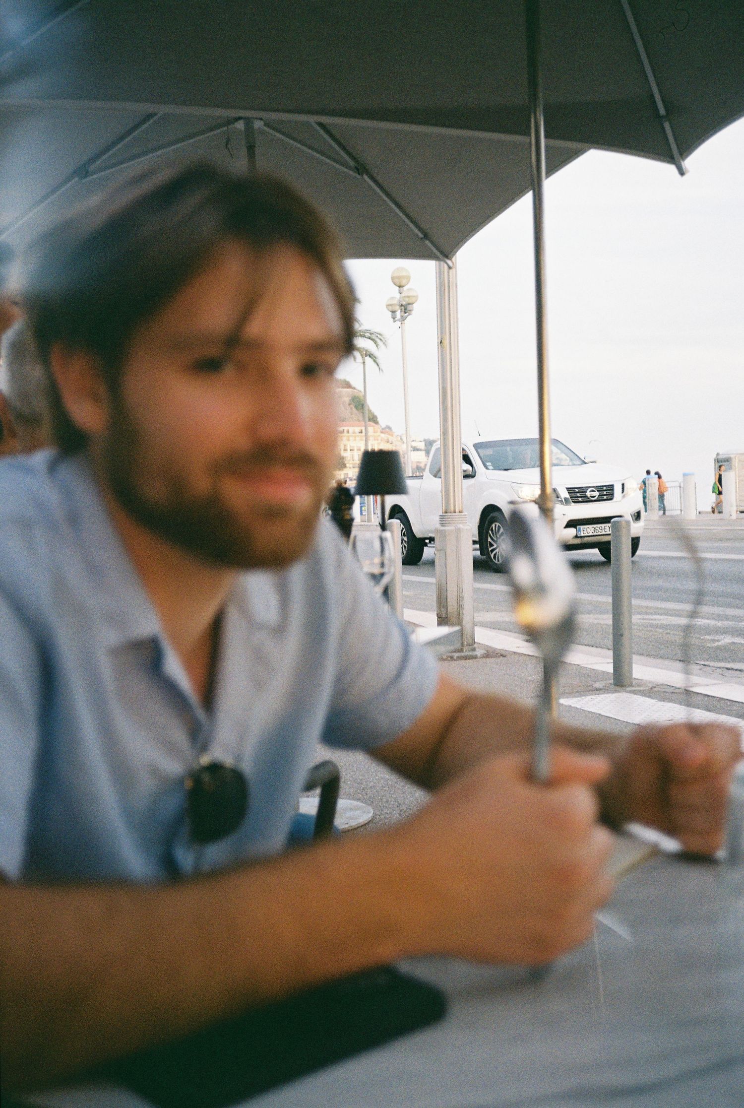 Bearded man sitting at an outdoor cafe table holding a wine glass under an umbrella.