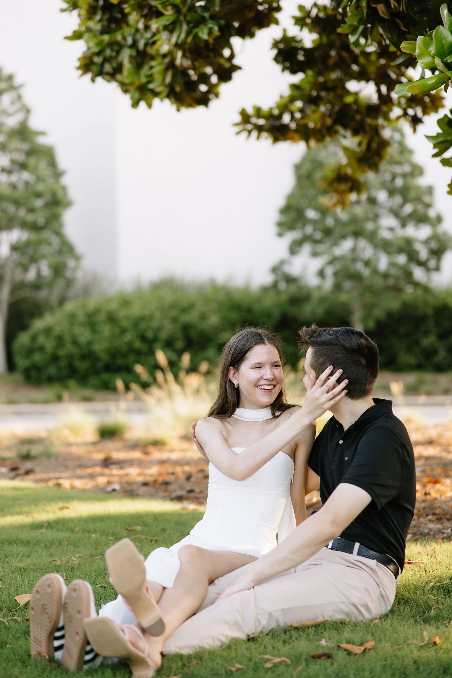 Couple sitting on grass laughing together under a tree during an outdoor engagement photo session.