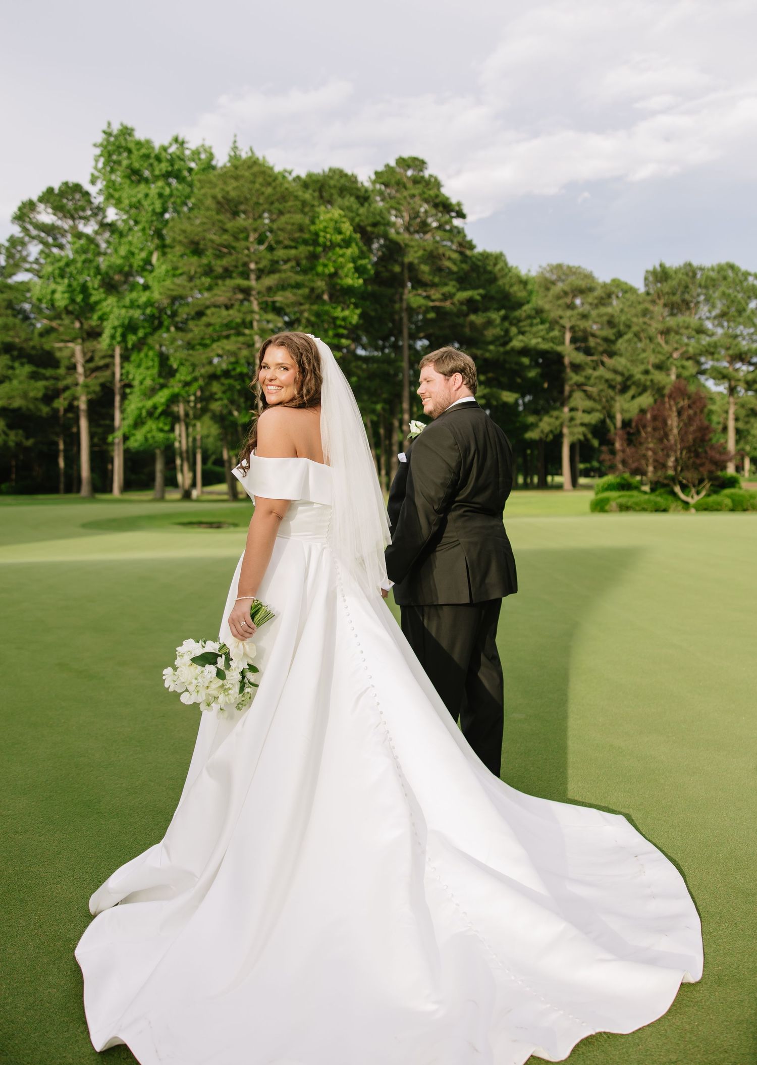 Bride in off-shoulder gown with cathedral train and groom in black tux on a golf course green.
