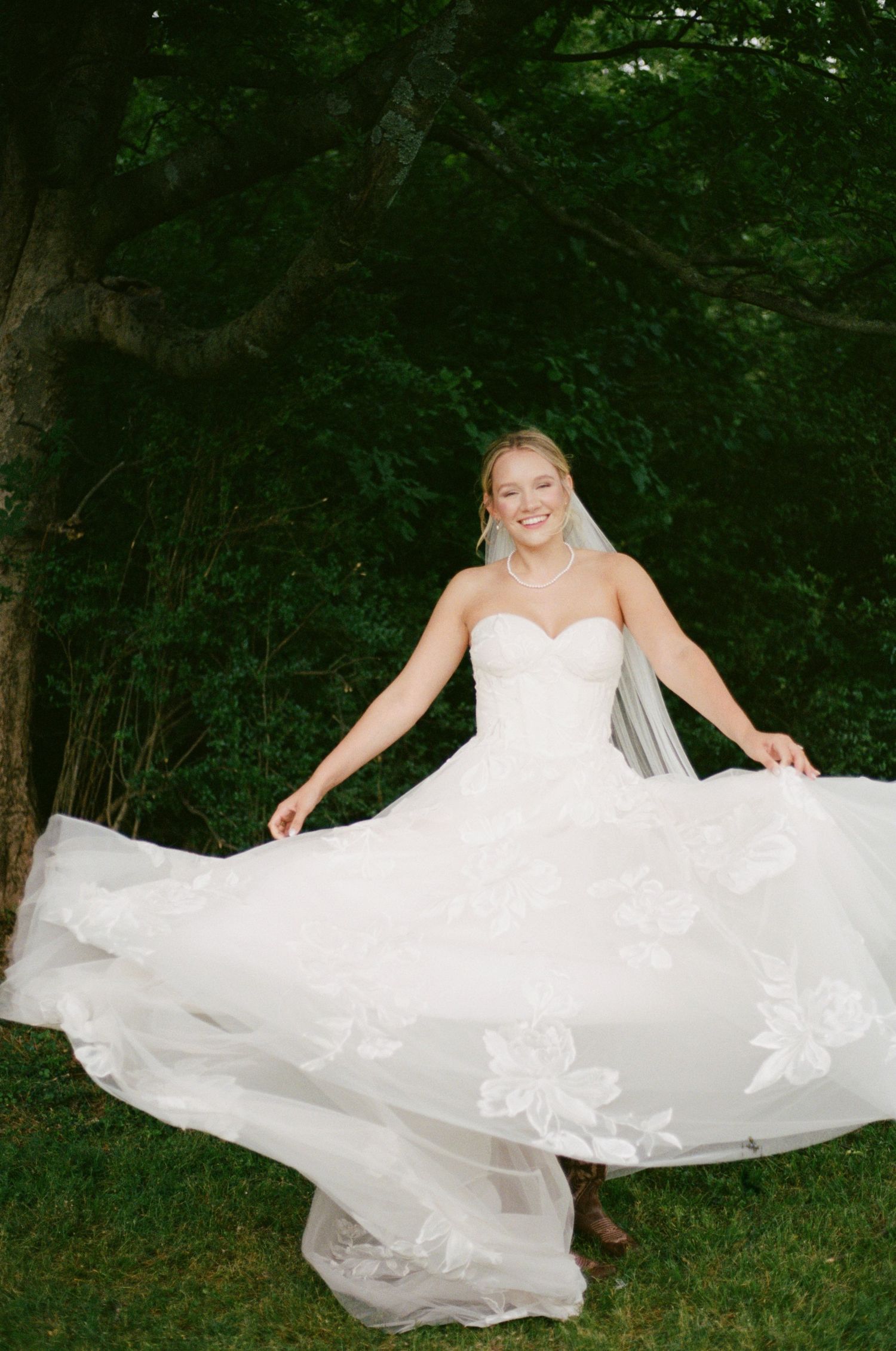 Radiant bride twirling in a strapless white floral lace wedding gown with veil in a lush garden setting.