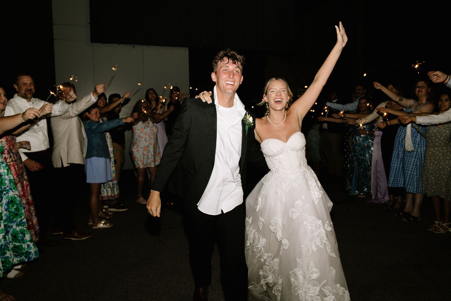 Joyful bride and groom exit wedding reception through sparkler tunnel at night celebration.