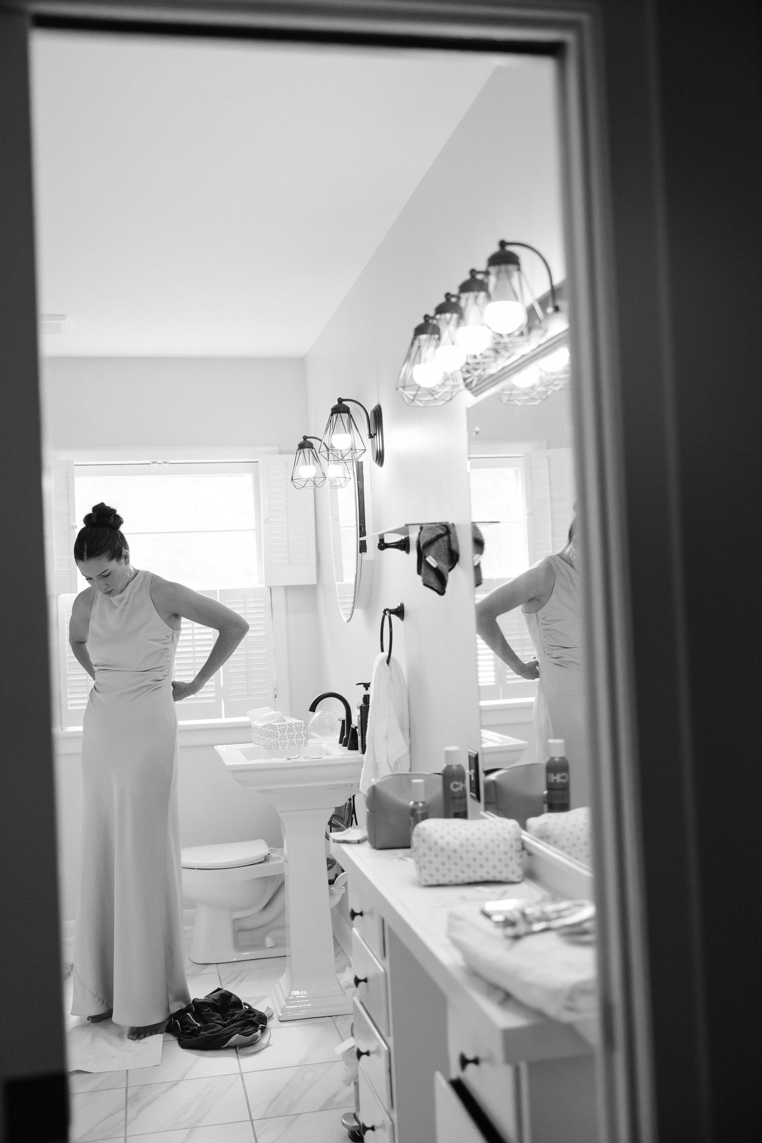 Black and white photo of a bridesmaid adjusting her dress in a bathroom mirror reflection.