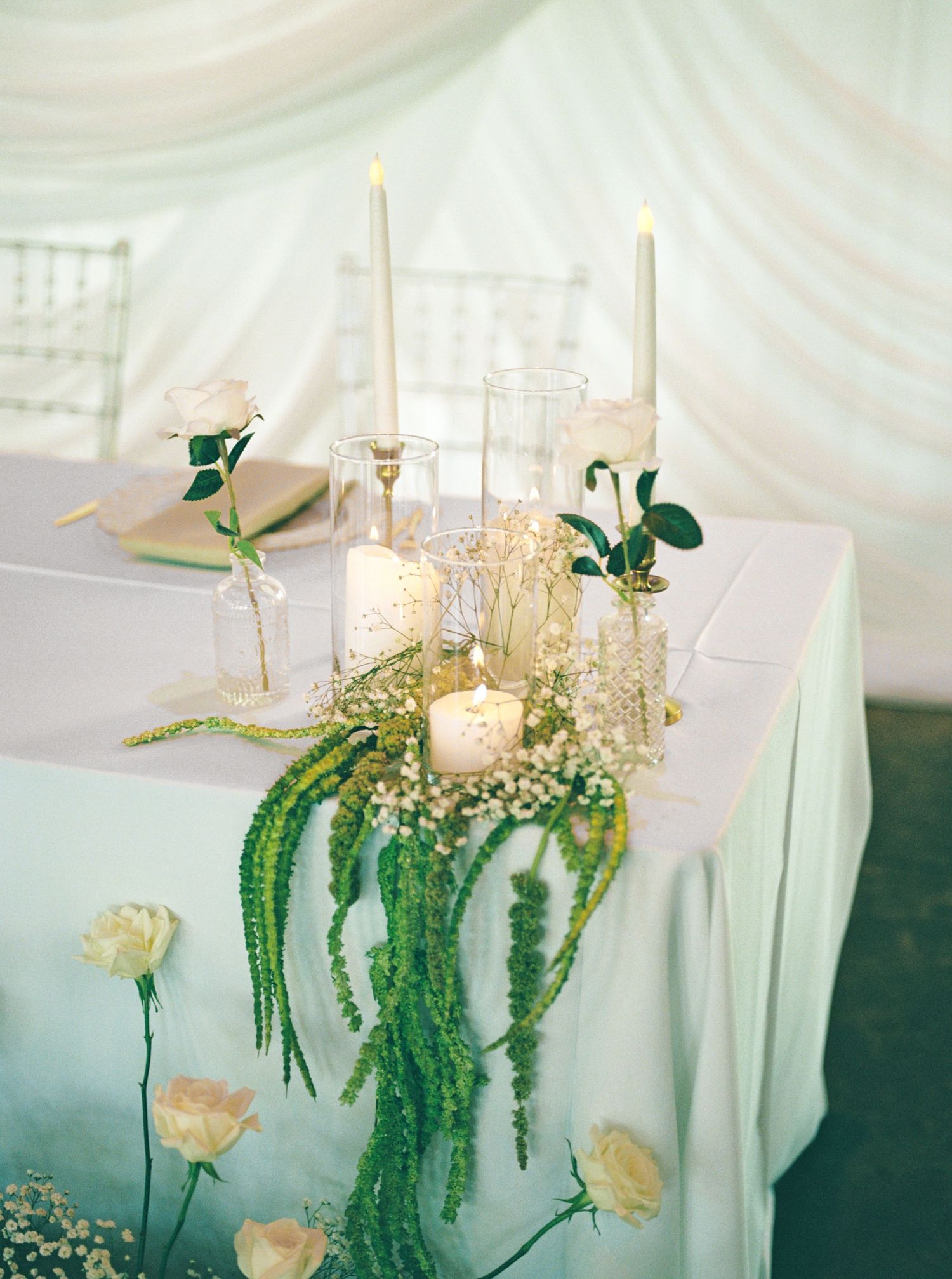 Elegant wedding reception table with white roses, baby's breath, greenery garland, candles, and tall taper candles under draped fabric.