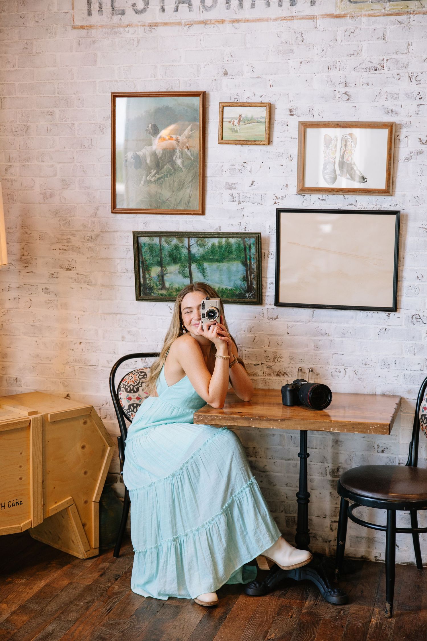 Woman in light blue maxi dress sitting at wooden table in vintage cafe, holding camera, smiling, with framed art on brick wall.