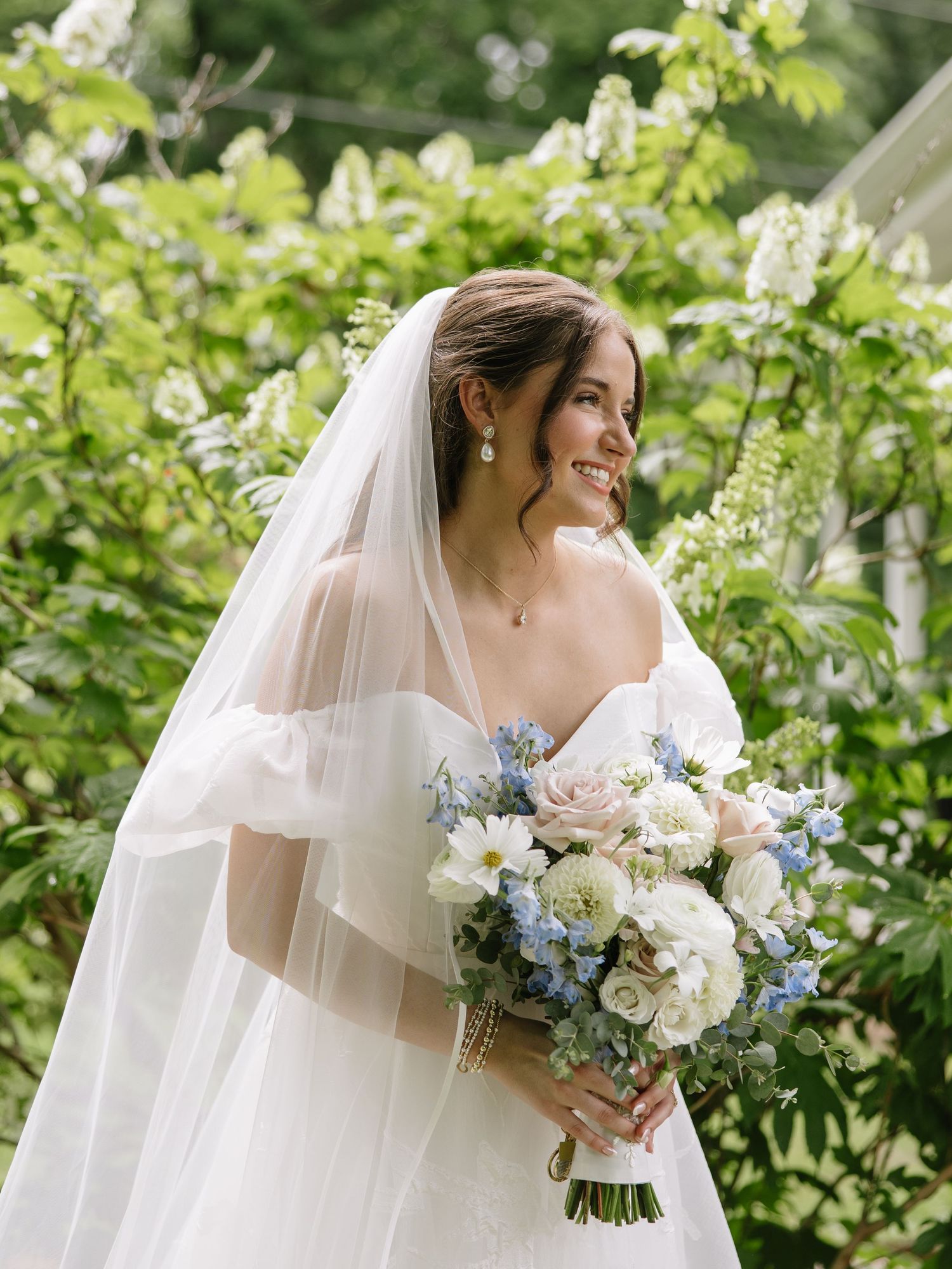 Smiling bride in off-shoulder gown and veil holds blue and white bouquet outdoors among lush green foliage.