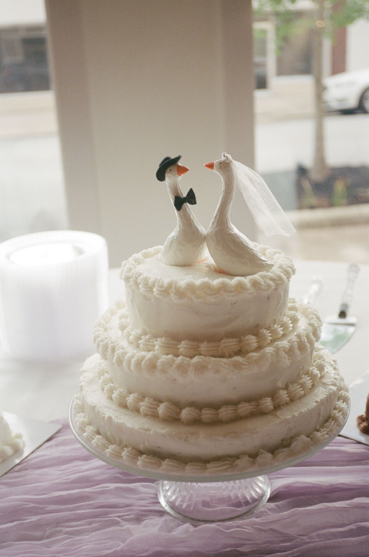 Three-tier white wedding cake with swan bride and groom cake toppers on a glass pedestal stand.