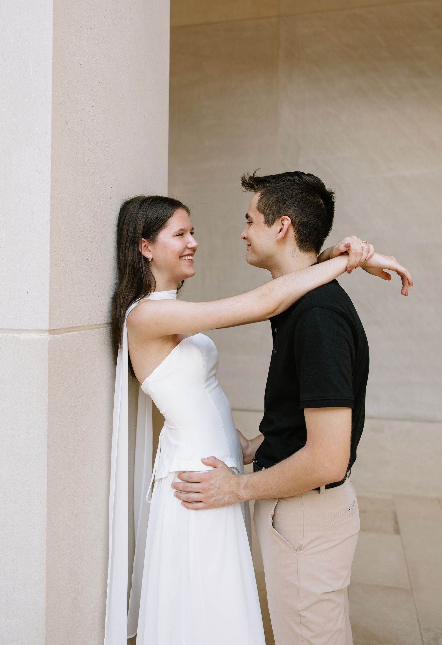 Couple sharing a romantic moment by stone columns, woman in white dress smiling as man in black shirt embraces her.
