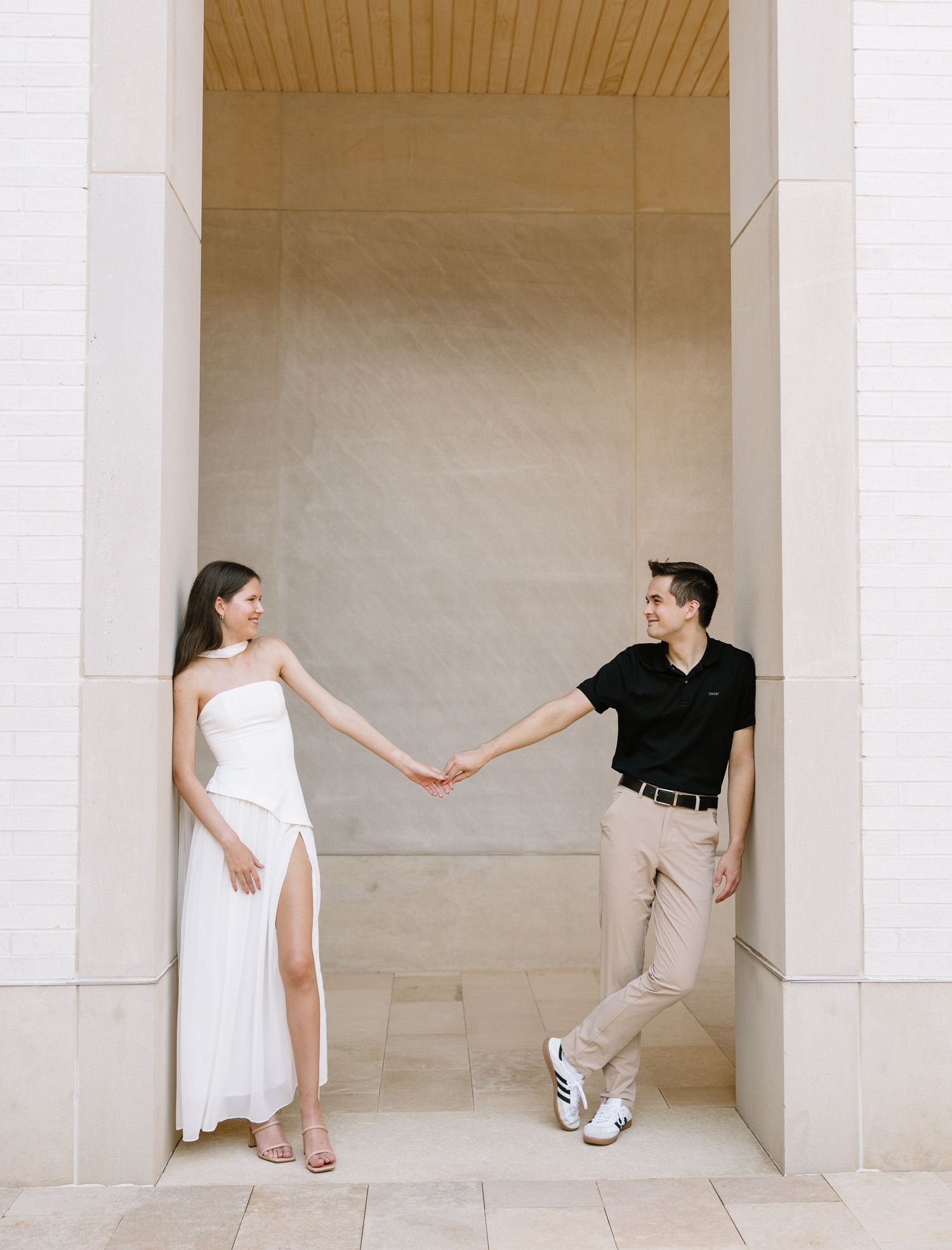 Couple holding hands in elegant white dress and black polo shirt posing between marble columns for engagement photos.