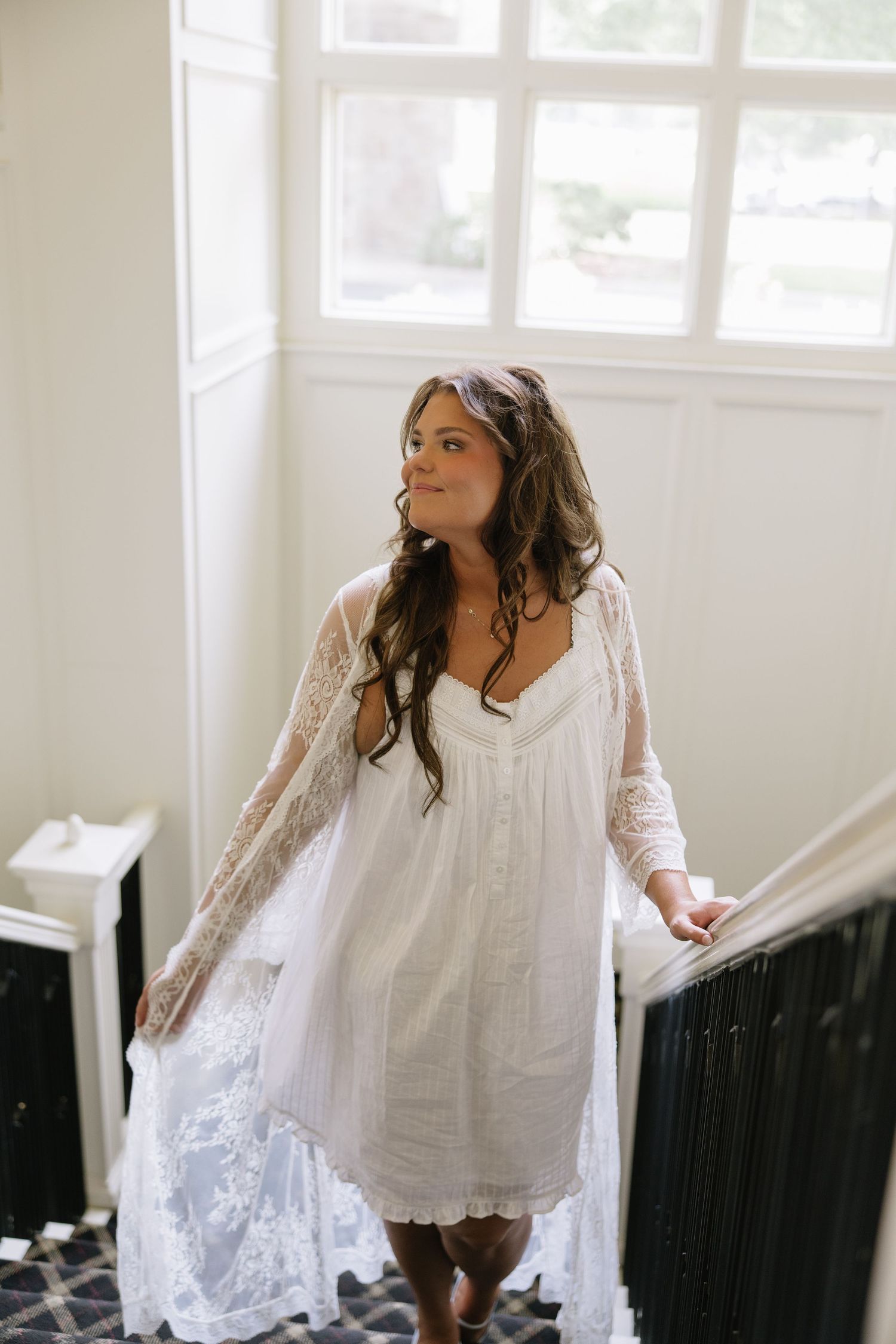 Bride in white lace robe and dress standing on staircase near window with flowing brunette hair.