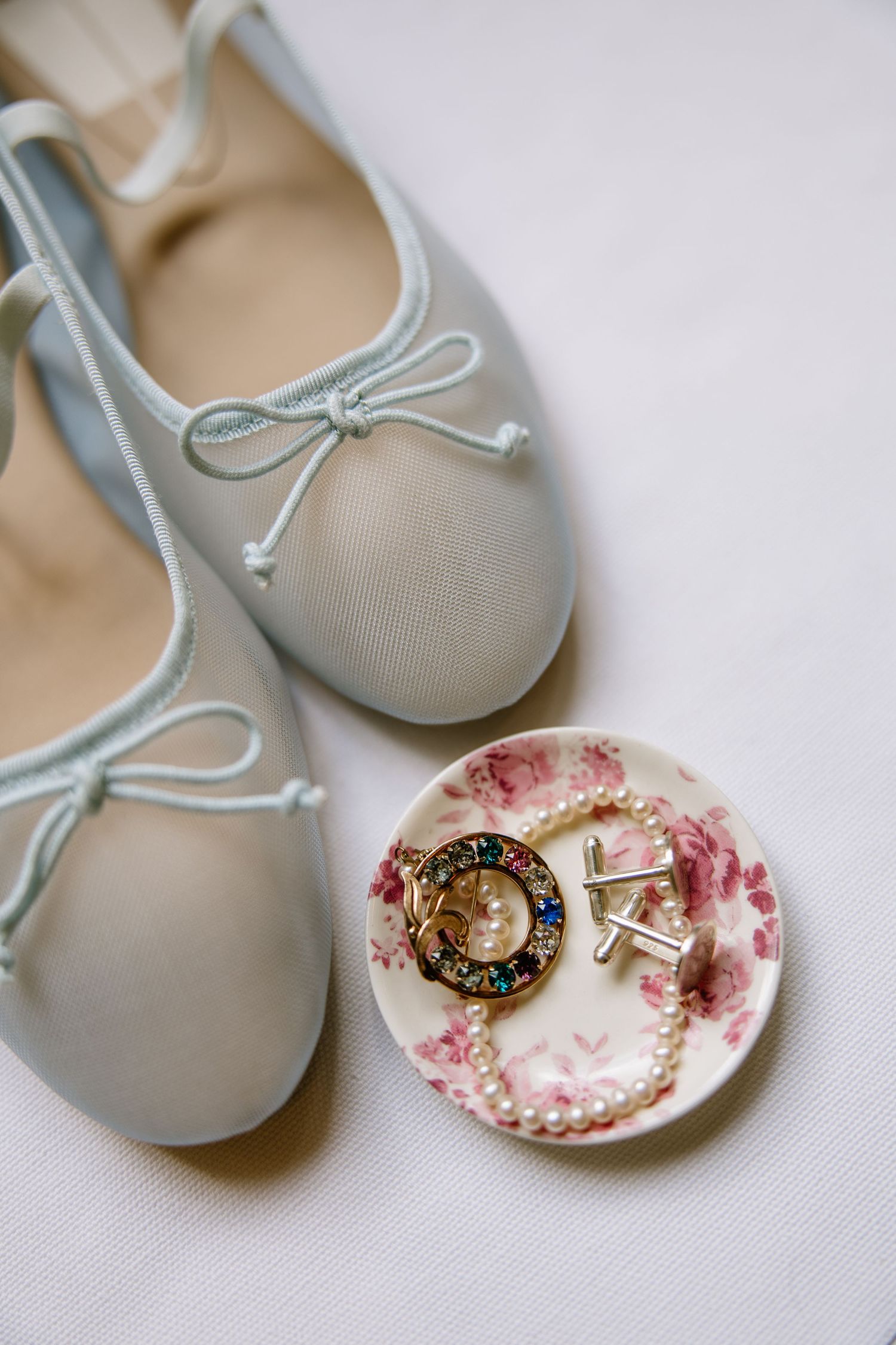 Light blue ballet flats with bow detail beside a floral dish holding pearl bracelet, gold brooch, and cufflinks.