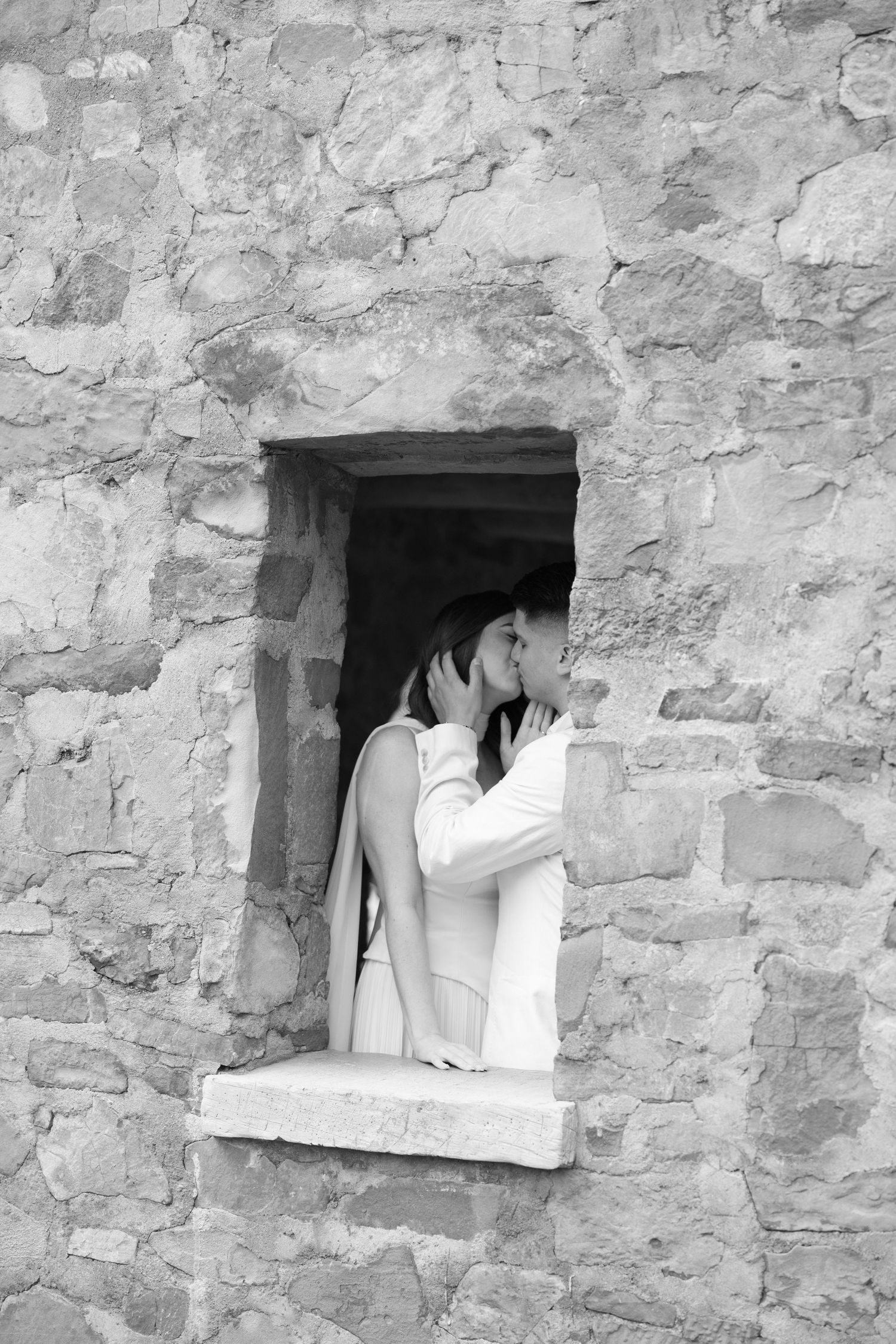 Black and white photo of a woman in white jacket leaning out of a rustic stone wall window opening.