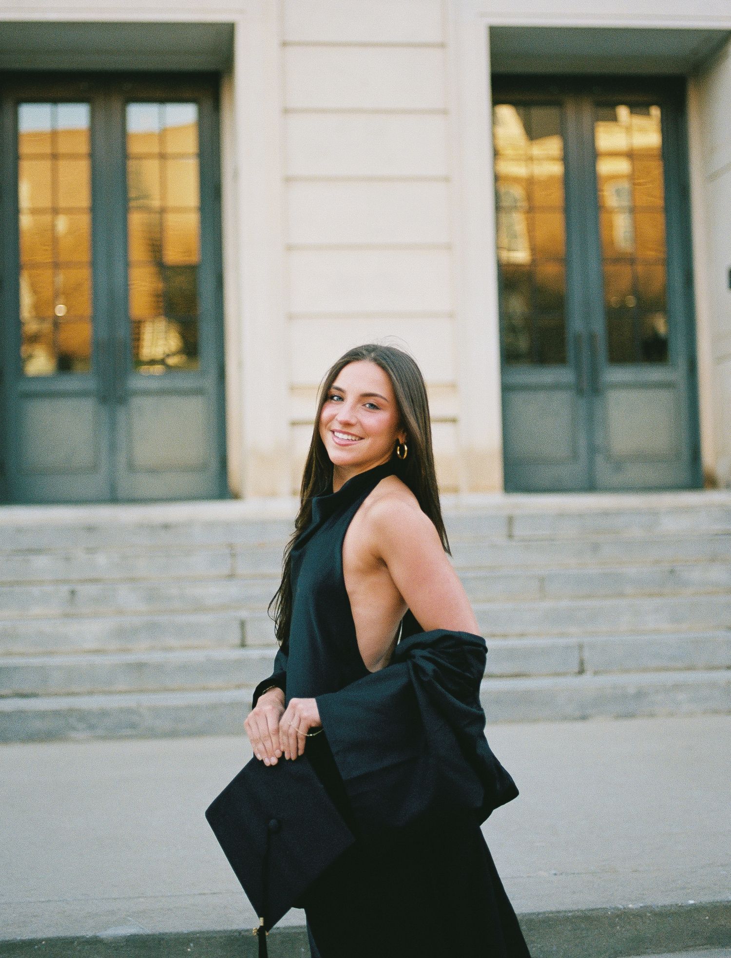 Stylish graduate in black dress carrying graduation gown over shoulder, smiling outside a classic building.