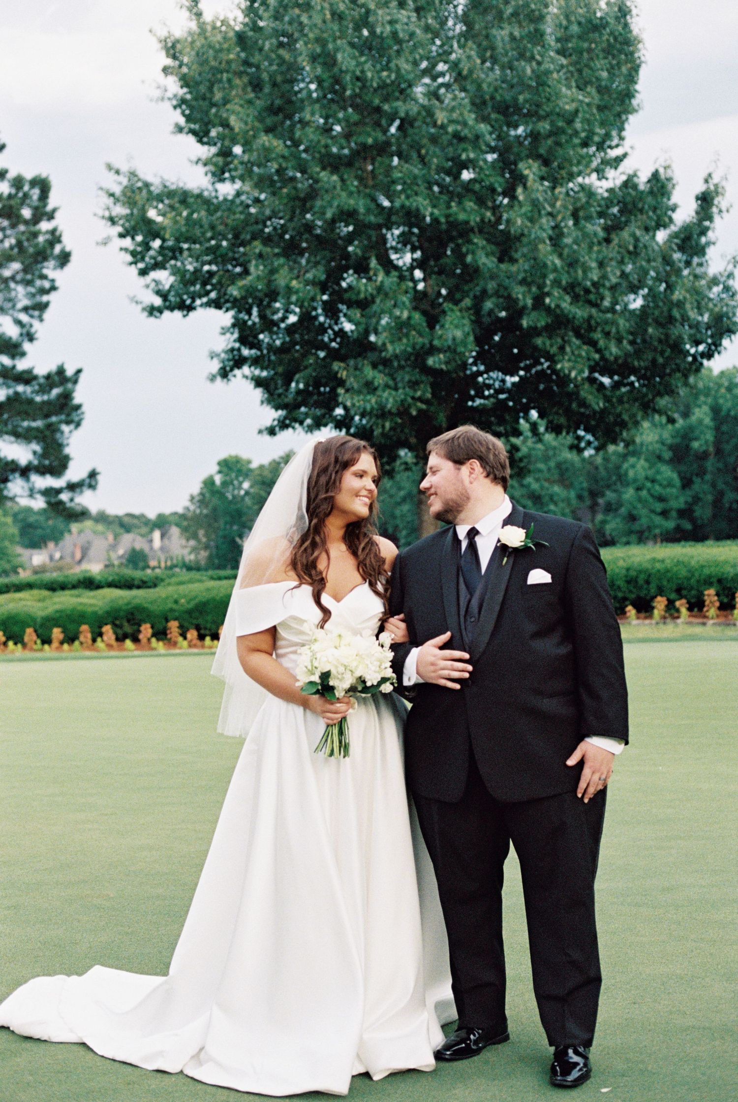 Bride and groom share a romantic moment on a green lawn, she holds white bouquet, he wears black tuxedo.
