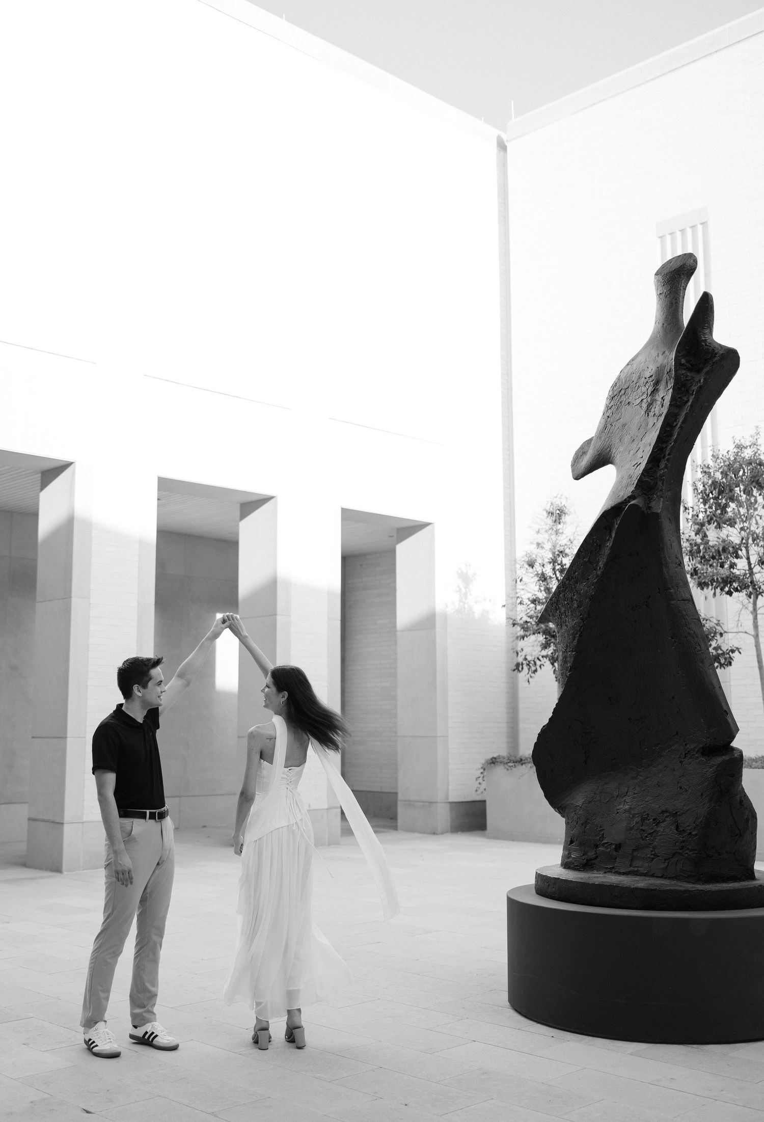 Black and white photo of a couple dancing near a large abstract sculpture in a modern architectural courtyard.