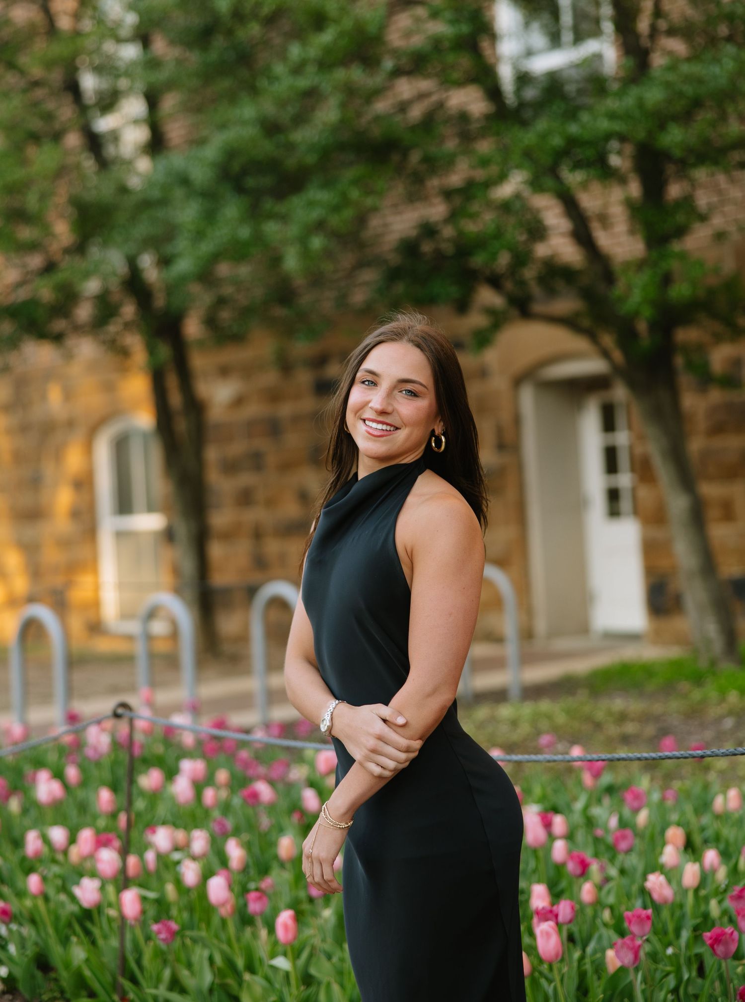 Smiling young woman in black sleeveless dress posing near pink tulips outside brick university building.
