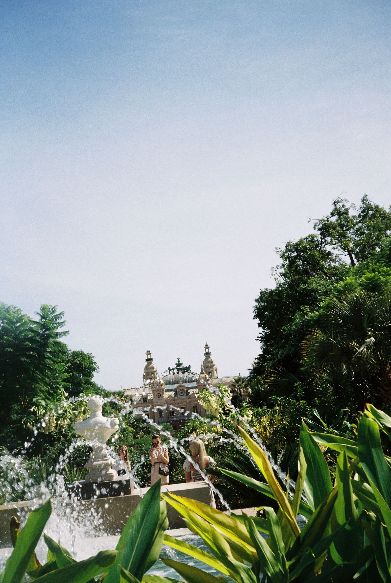 Ornate Baroque building with fountain and tropical plants under clear blue sky in Monaco.