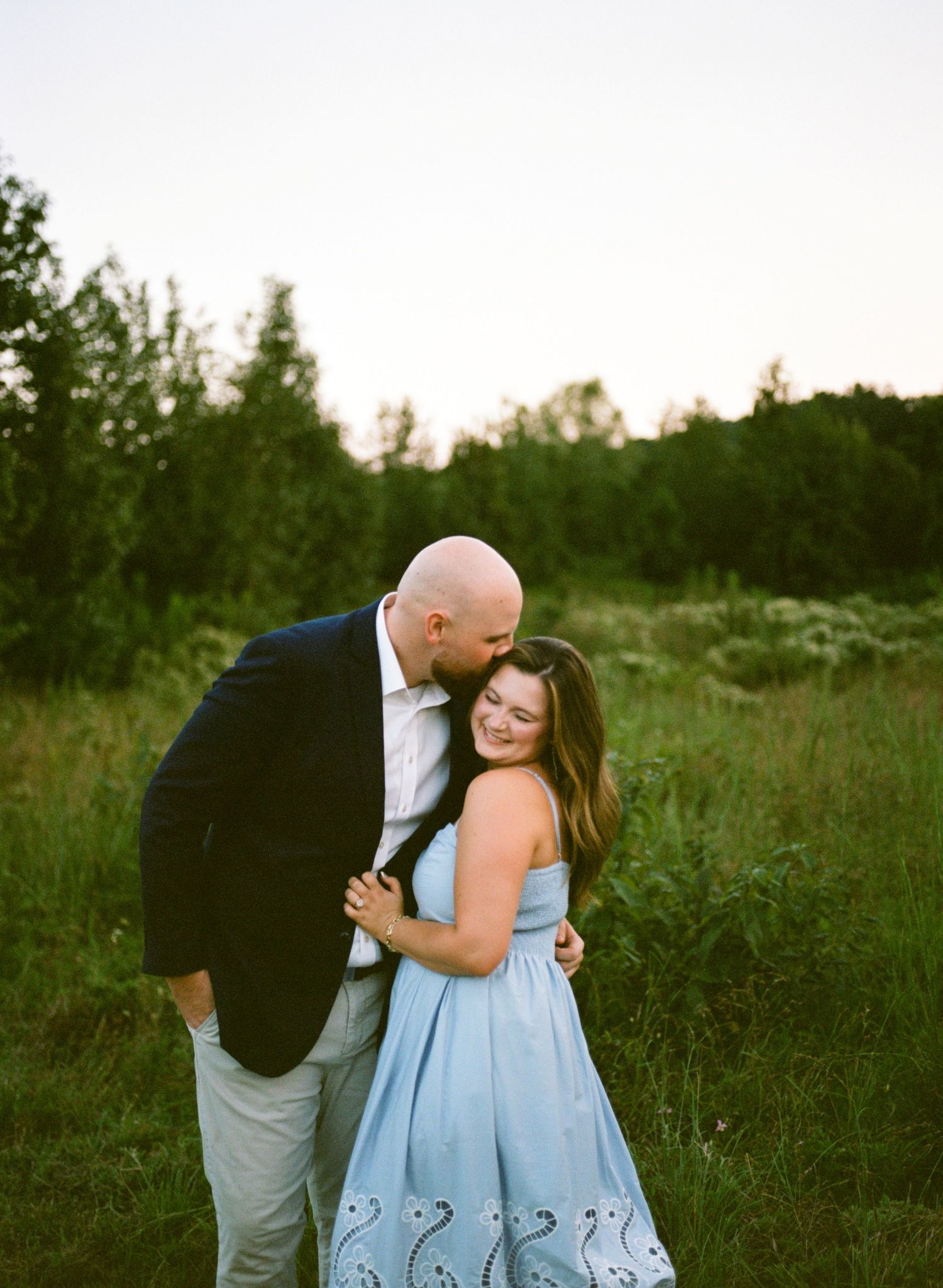A couple shares a tender embrace in a lush green field at golden hour, woman in blue dress smiling joyfully.