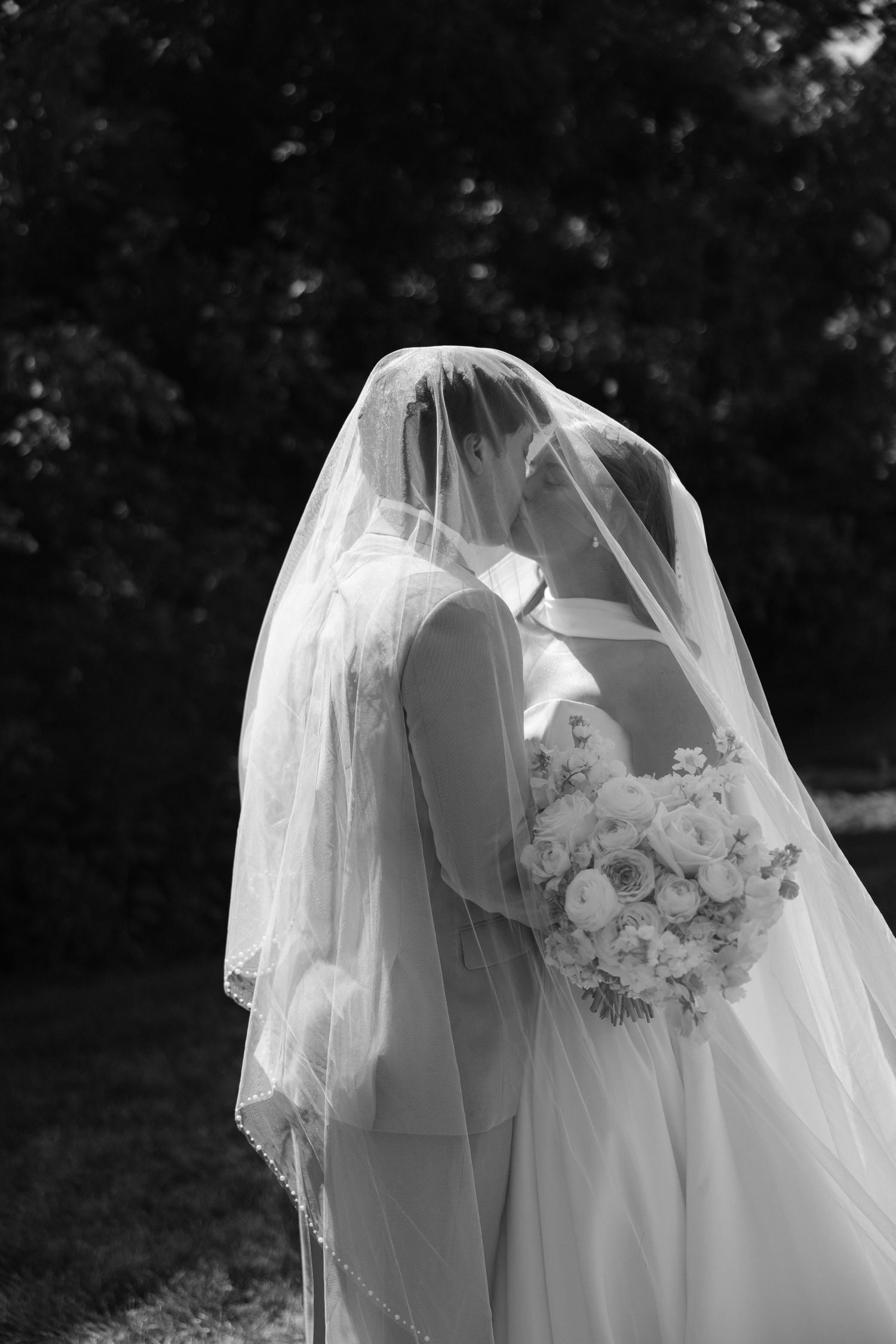 Black and white photo of two brides sharing a kiss under a wedding veil, holding a bouquet.