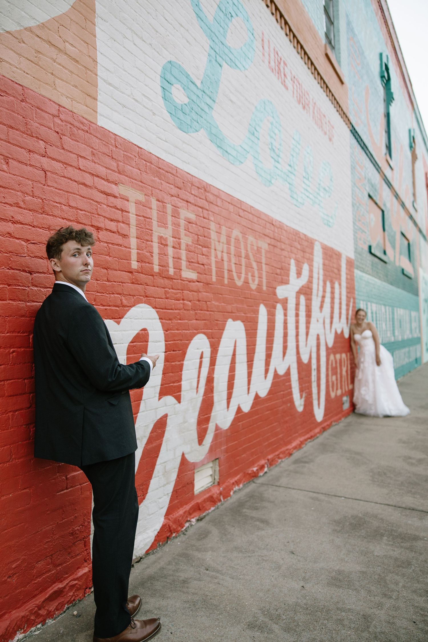 Groom in black suit leaning against colorful Love mural wall with bride in background.