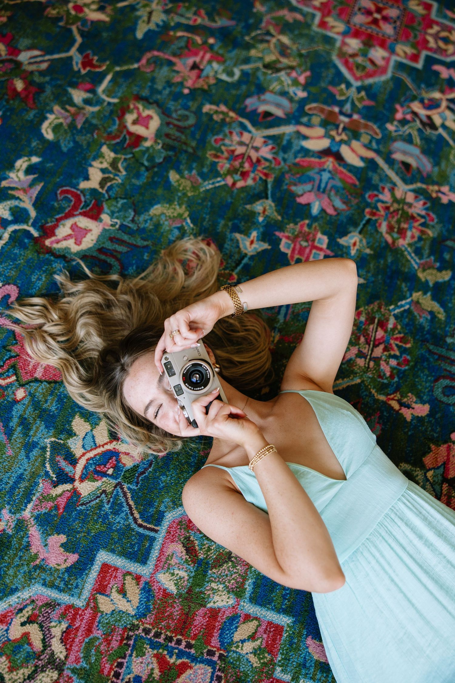 Young woman lying on a colorful floral rug, holding a vintage film camera up to her face while wearing a mint dress.