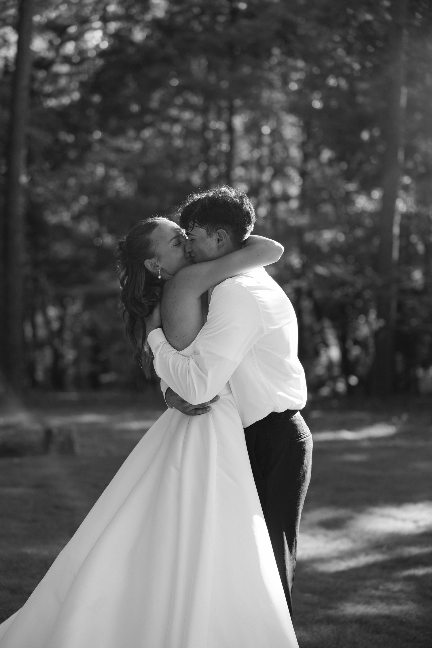Black and white photo of a bride and groom sharing a romantic kiss outdoors, surrounded by trees.
