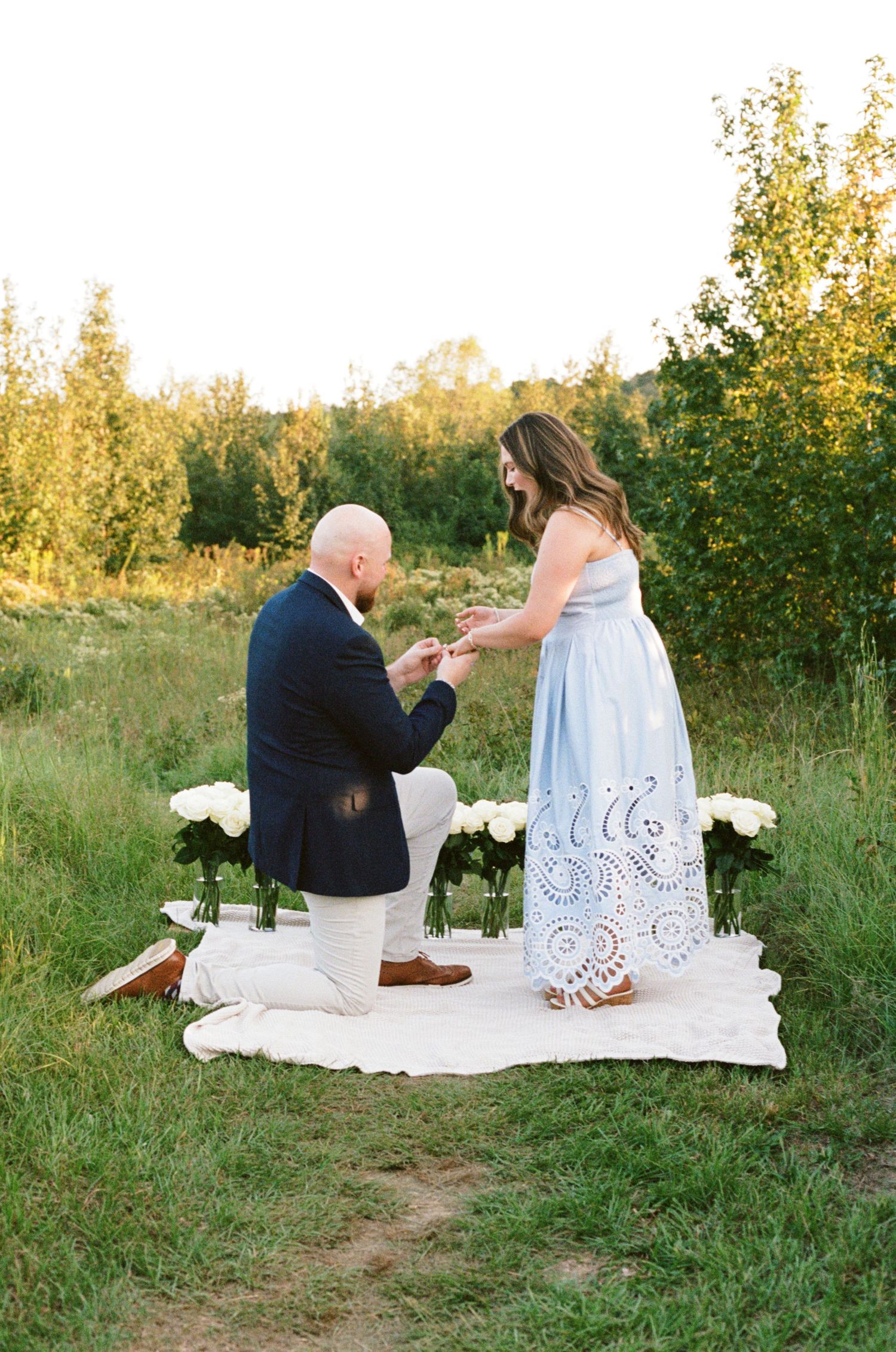 Man kneeling proposing to woman in blue lace dress on white blanket surrounded by white roses outdoors.