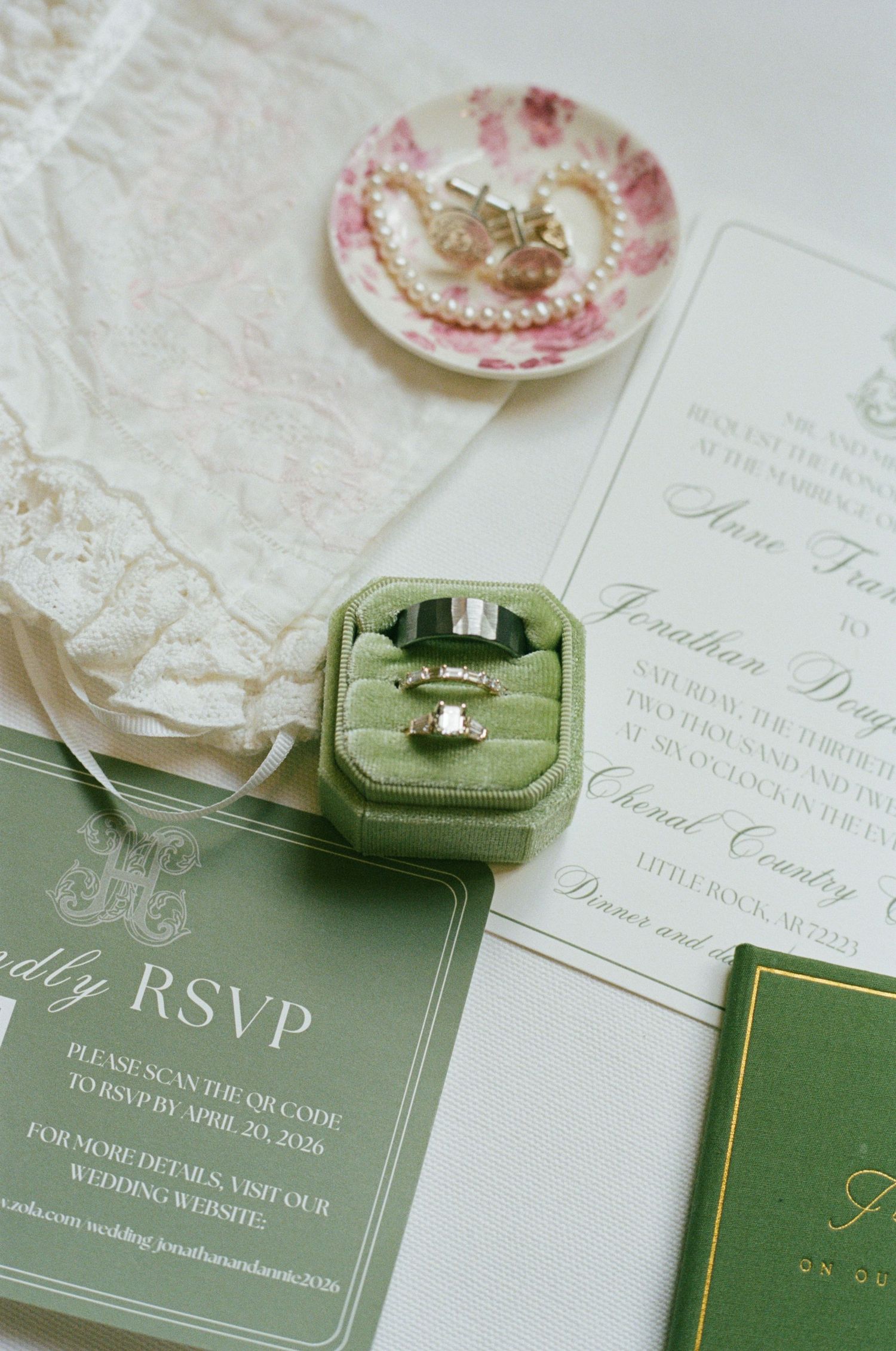 Green velvet ring box with wedding bands beside floral pearl dish, lace, and elegant green wedding invitations.