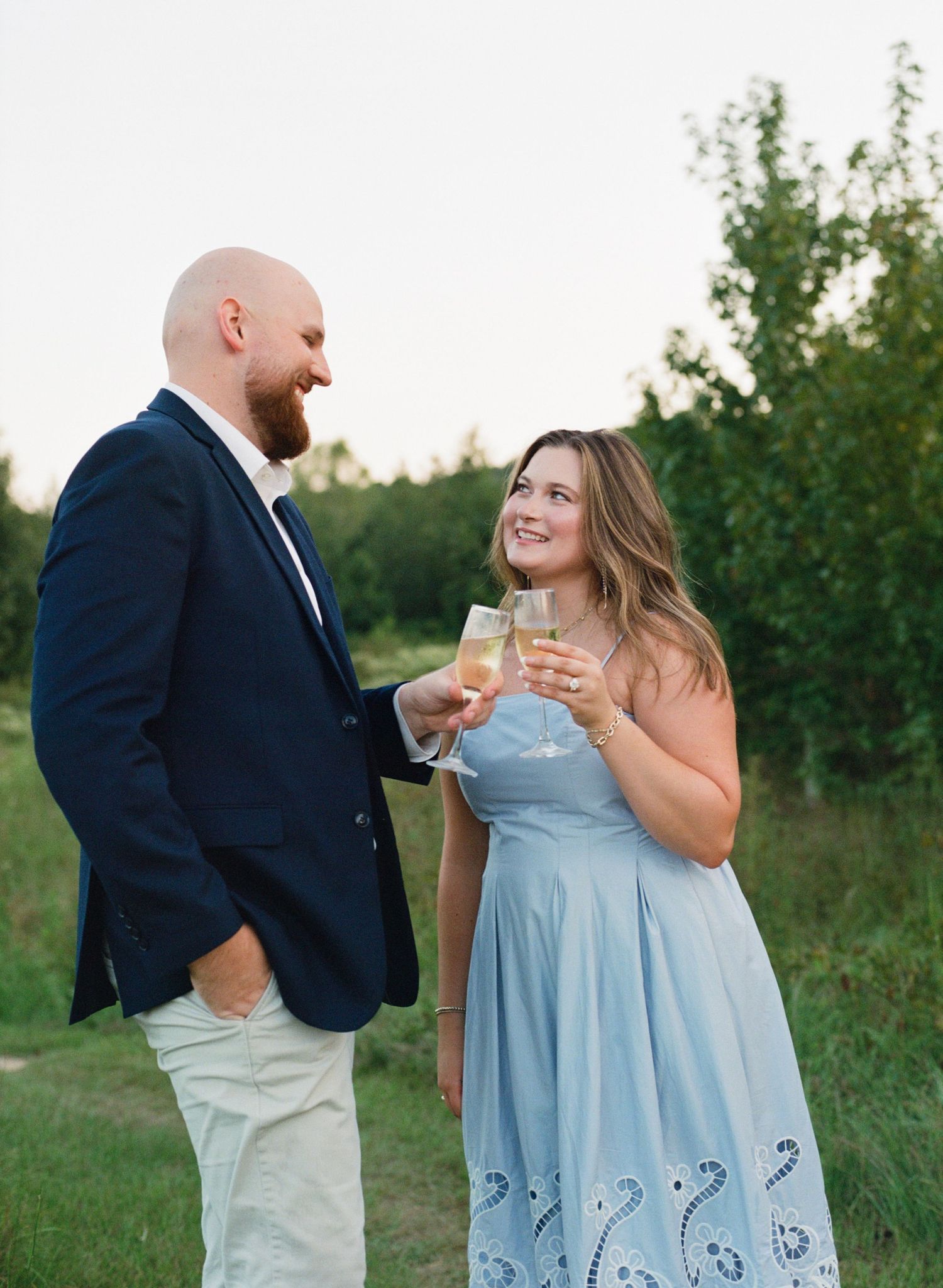 Couple toasting with champagne glasses outdoors at golden hour, man in navy blazer and woman in light blue dress.