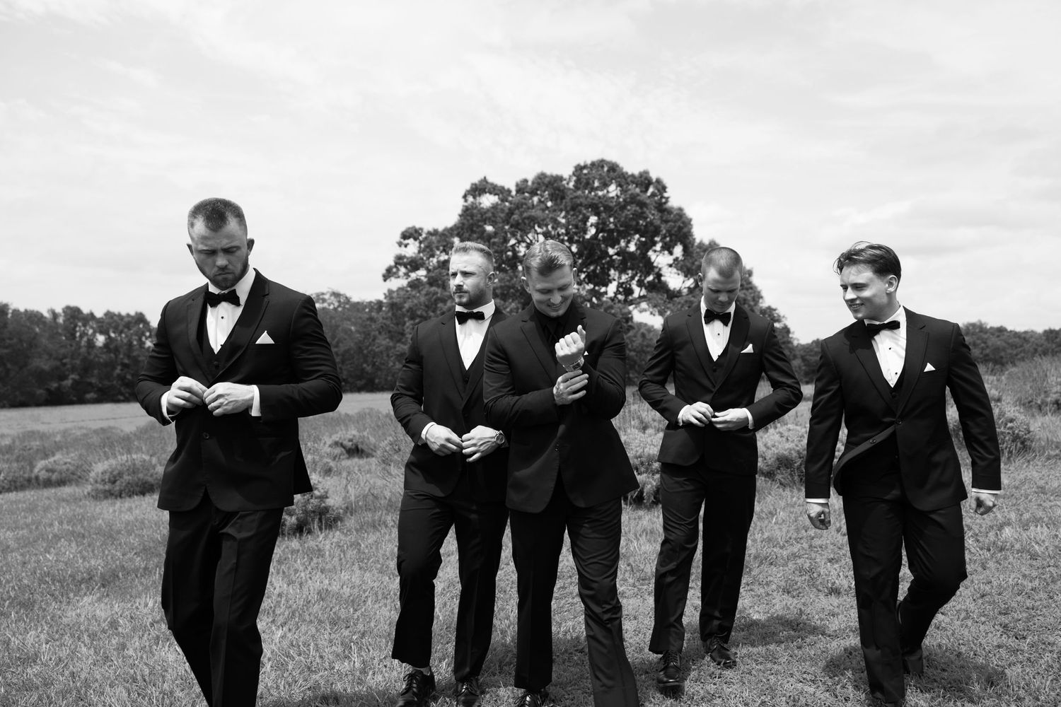 Black and white photo of five groomsmen in tuxedos walking outdoors in a field on a wedding day.