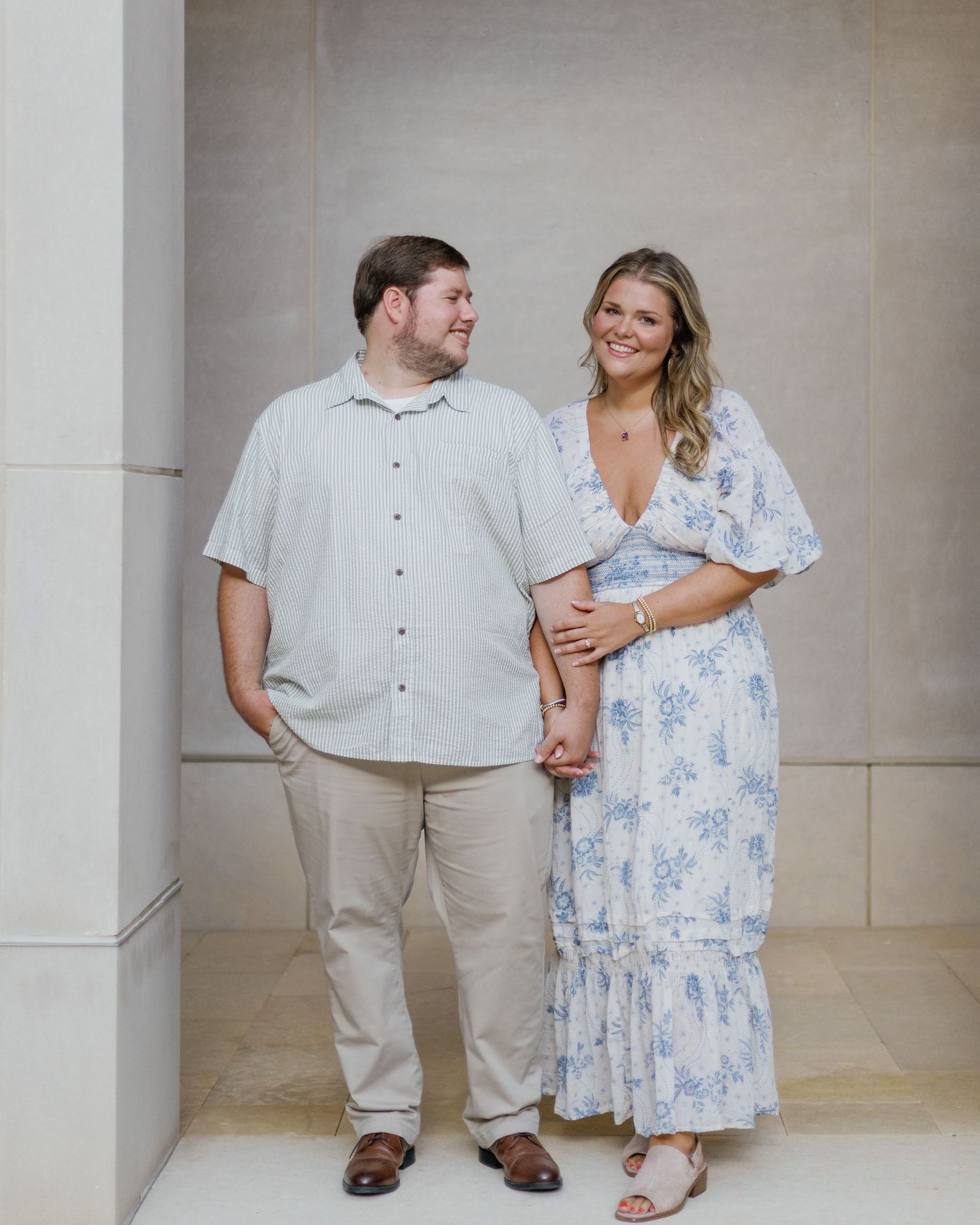 A smiling couple holds hands, man in light shirt and khakis, woman in blue floral maxi dress, posing outdoors.