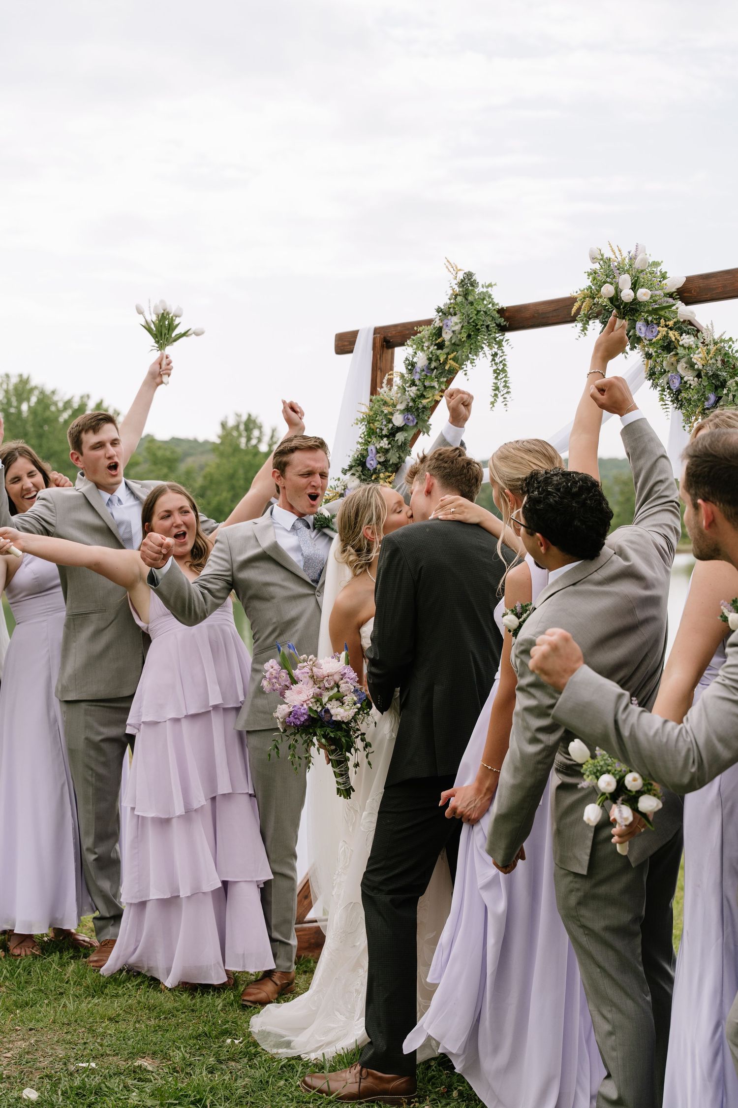 Wedding party cheers as bride and groom share first kiss under floral arch, bridesmaids in lavender gowns, groomsmen in gray suits.