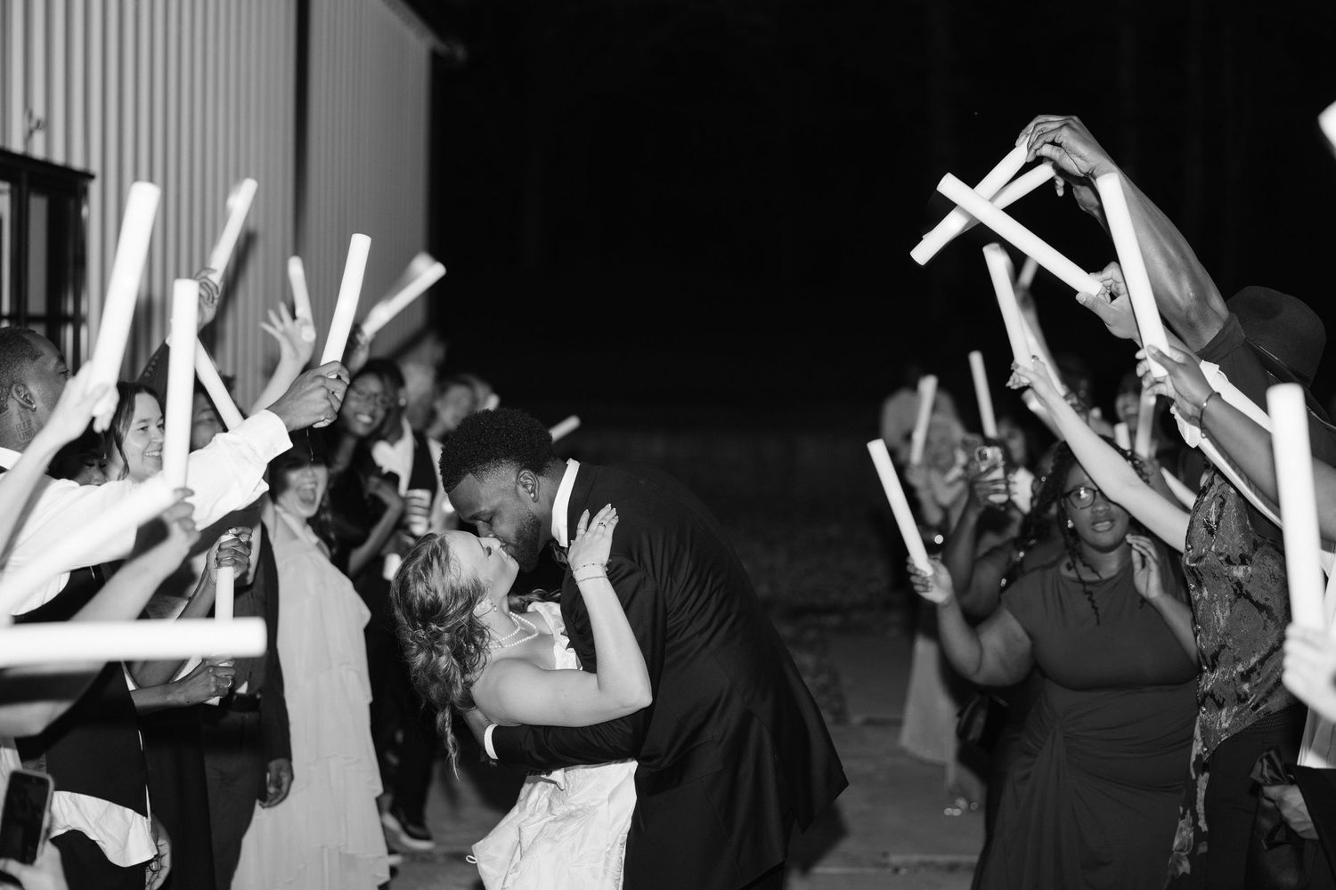 Black and white photo of newlyweds kissing under an arch of raised glow sticks held by wedding guests.