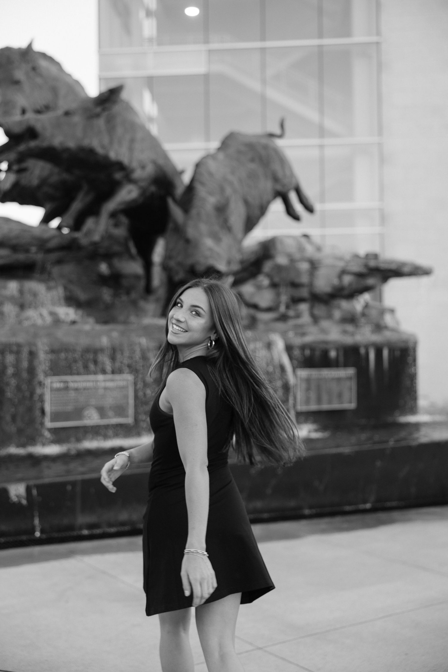 Smiling young woman in black dress posing in front of wild boar sculpture fountain in black and white.