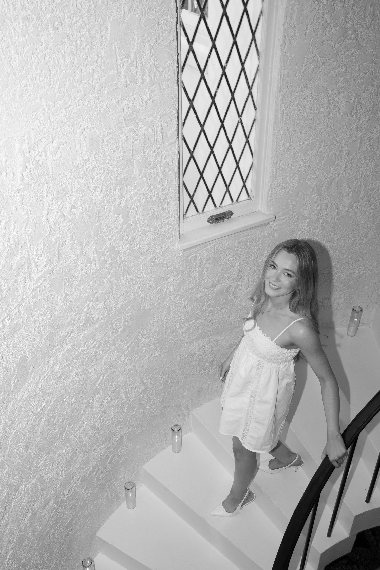 Black and white photo of a young blonde woman in a white dress on a spiral staircase.