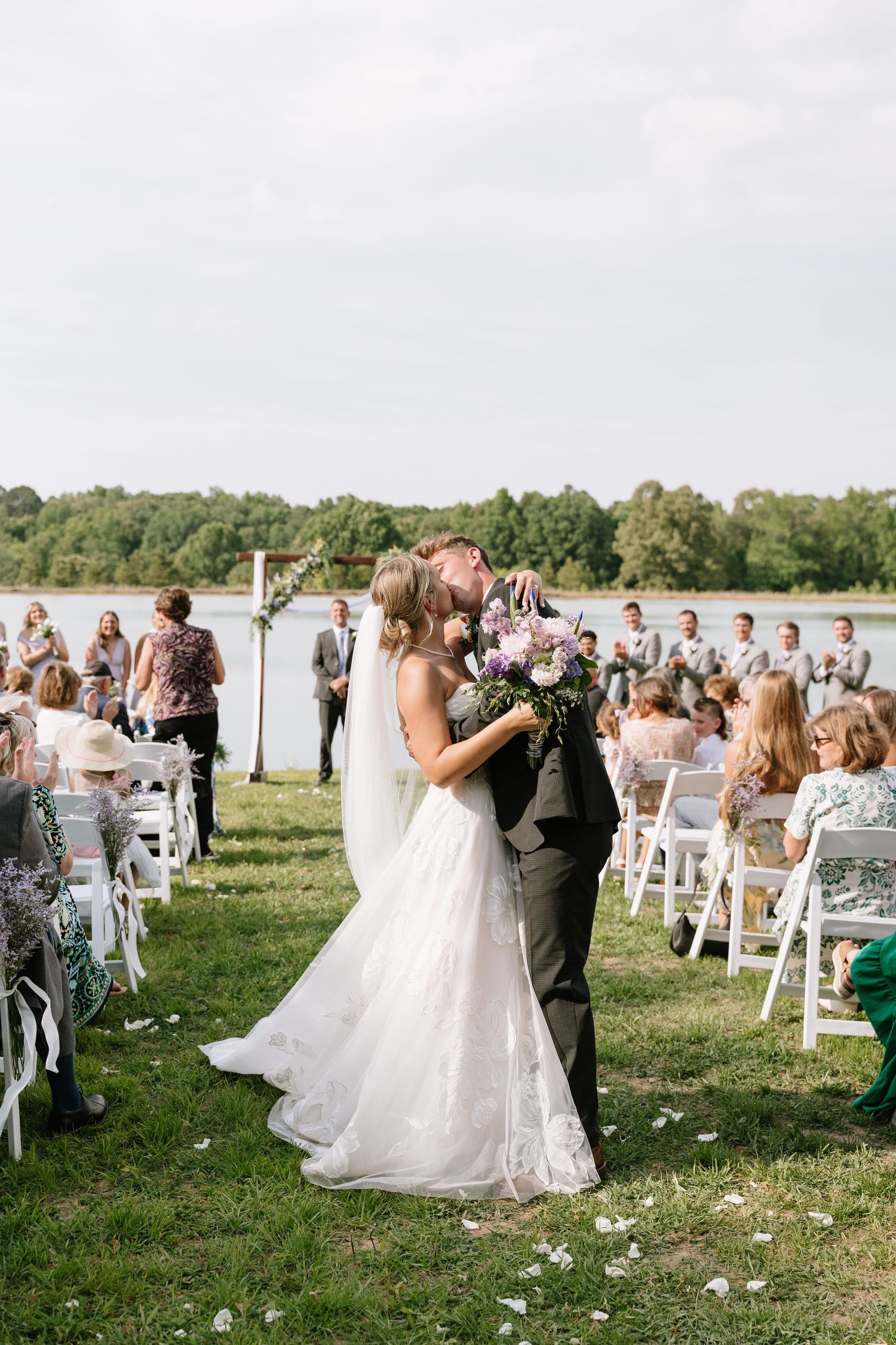 Bride and groom share first kiss at outdoor lakeside wedding ceremony surrounded by seated guests.