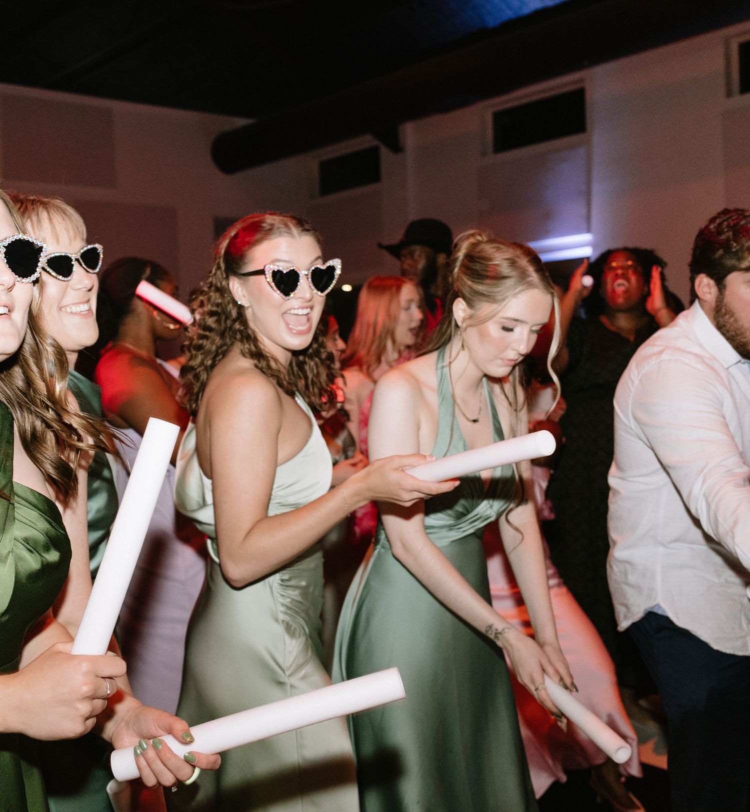 Bridesmaids in sage green dresses dancing with glow sticks at wedding reception.