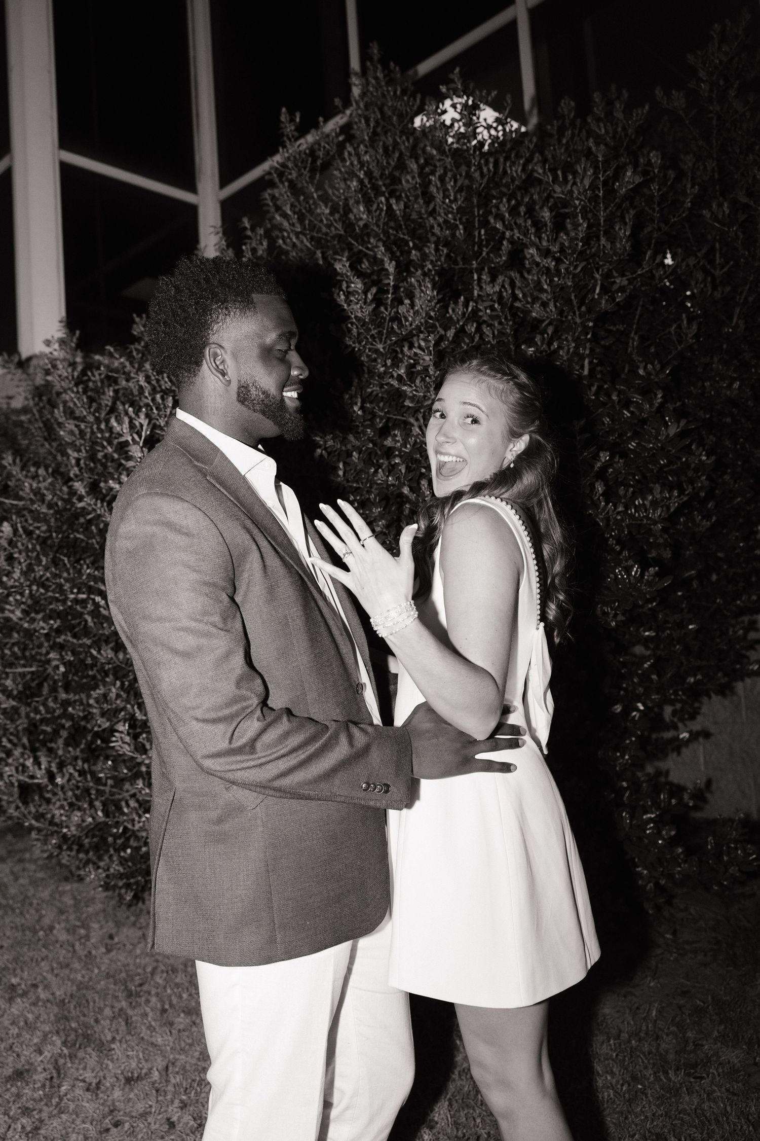 Couple smiling and showing off engagement ring outdoors at night in black and white photo.