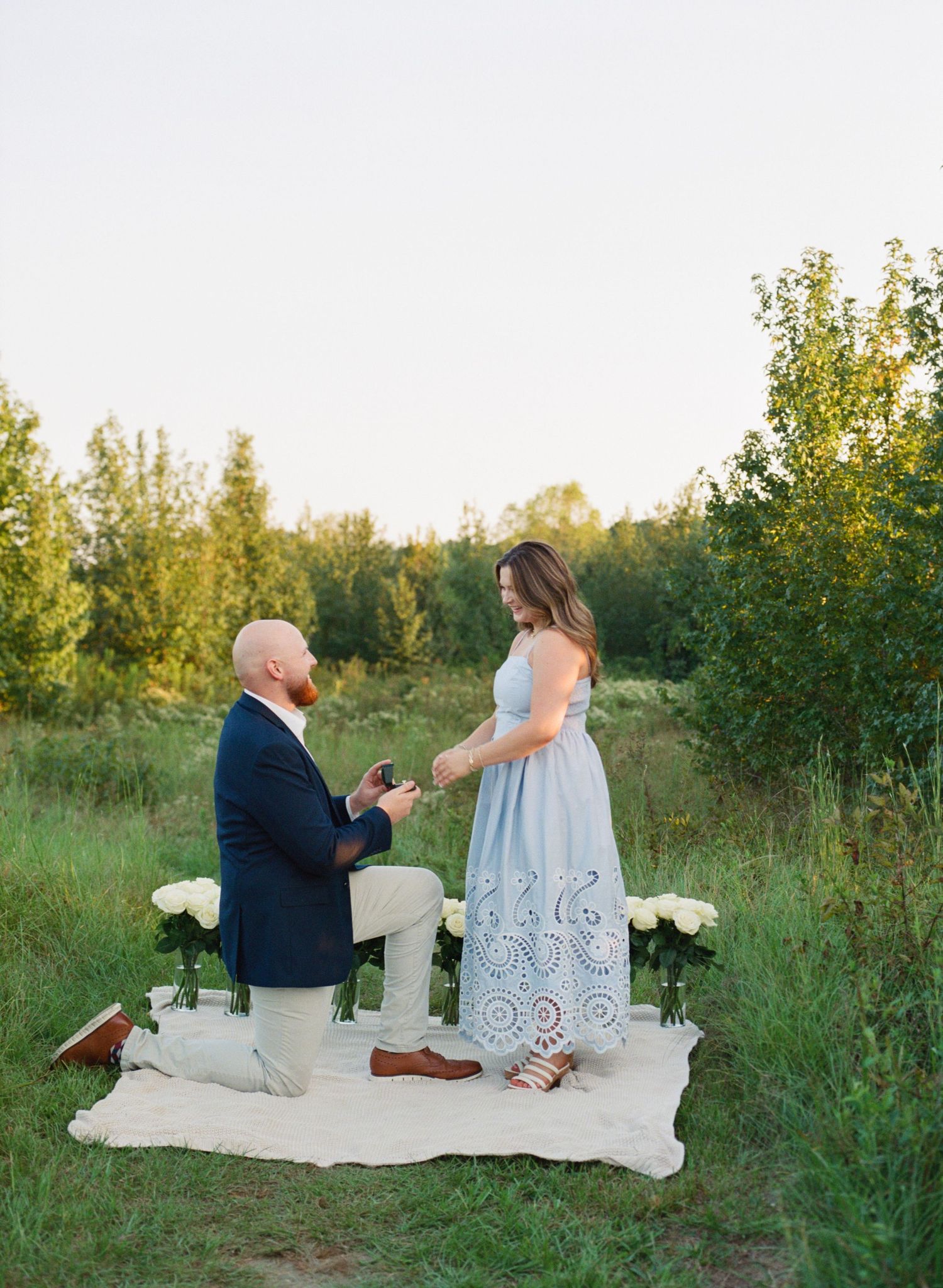 Man kneeling on blanket proposing to woman in blue dress outdoors at sunset with white flowers nearby.