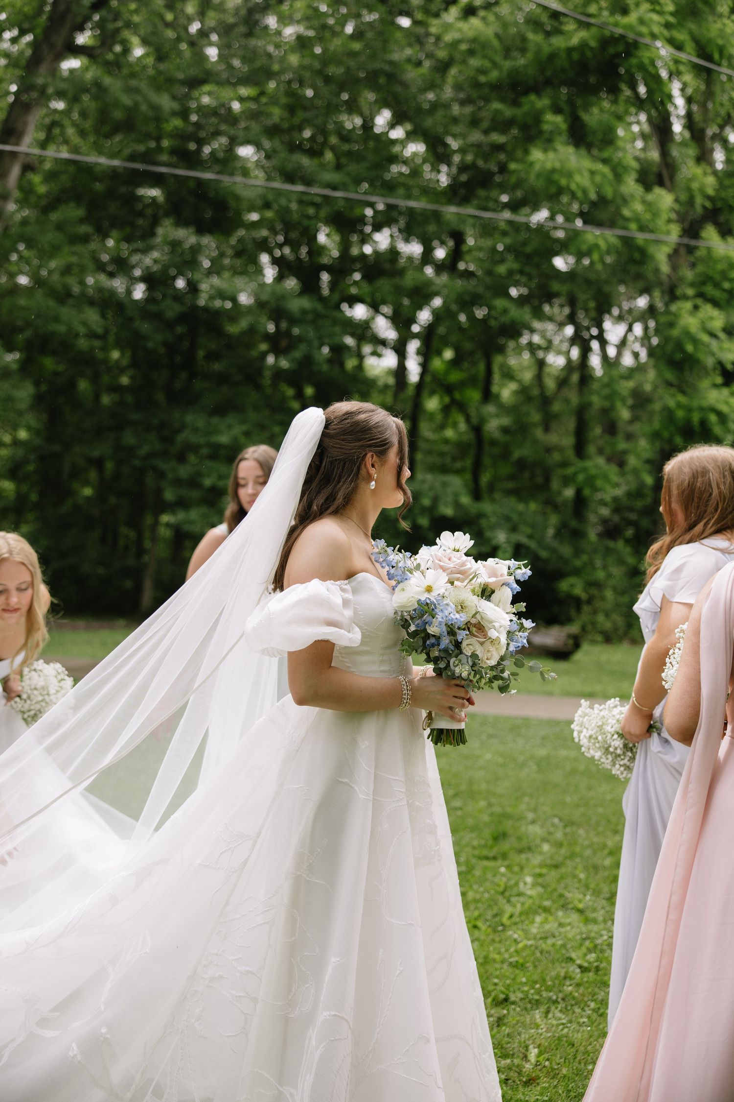 Bride in white gown with veil holding blue and white bouquet, standing with bridesmaids outdoors among lush green trees.