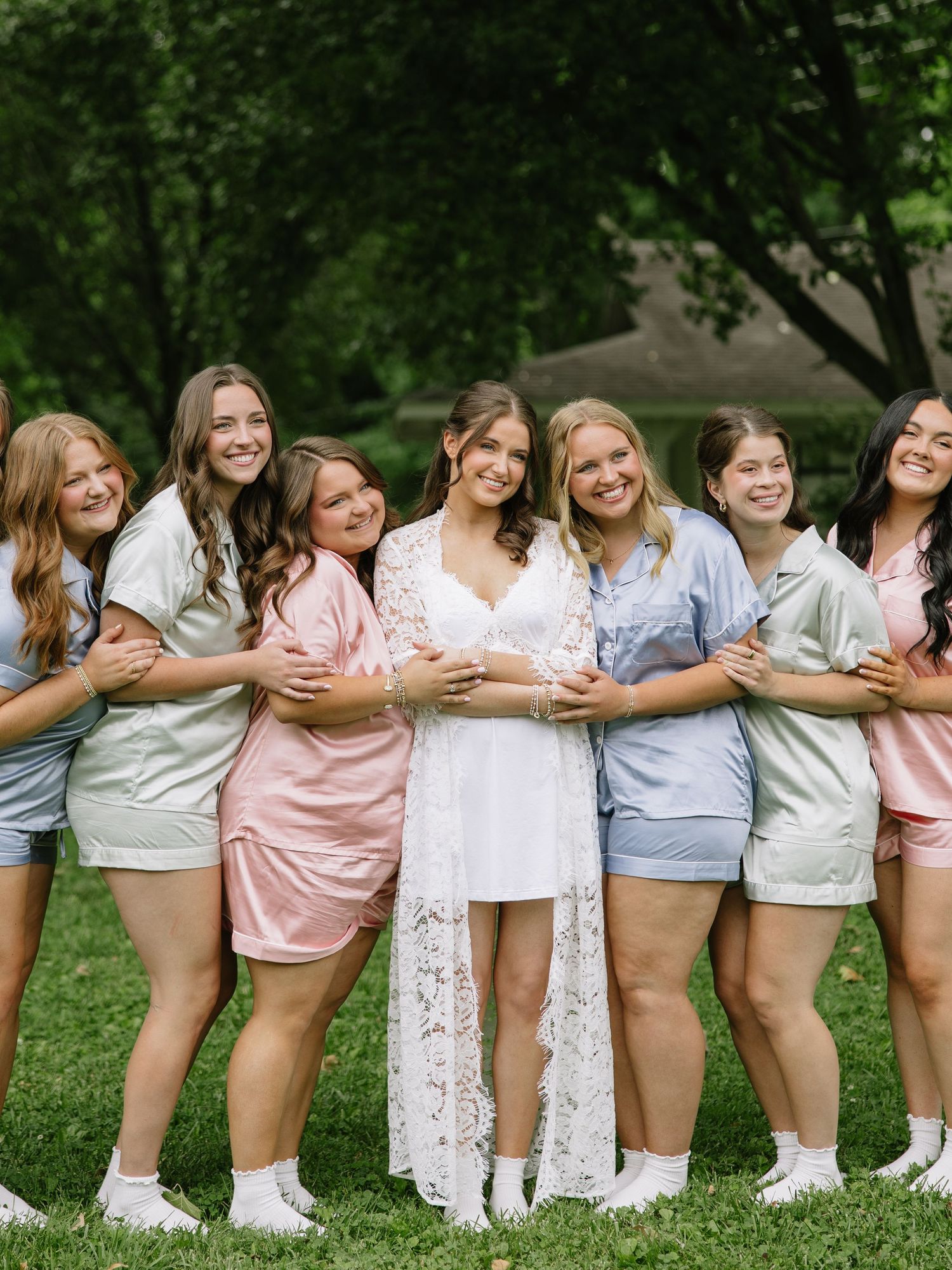 Bride in white lace robe surrounded by bridesmaids in colorful satin pajamas posing outdoors on green grass.