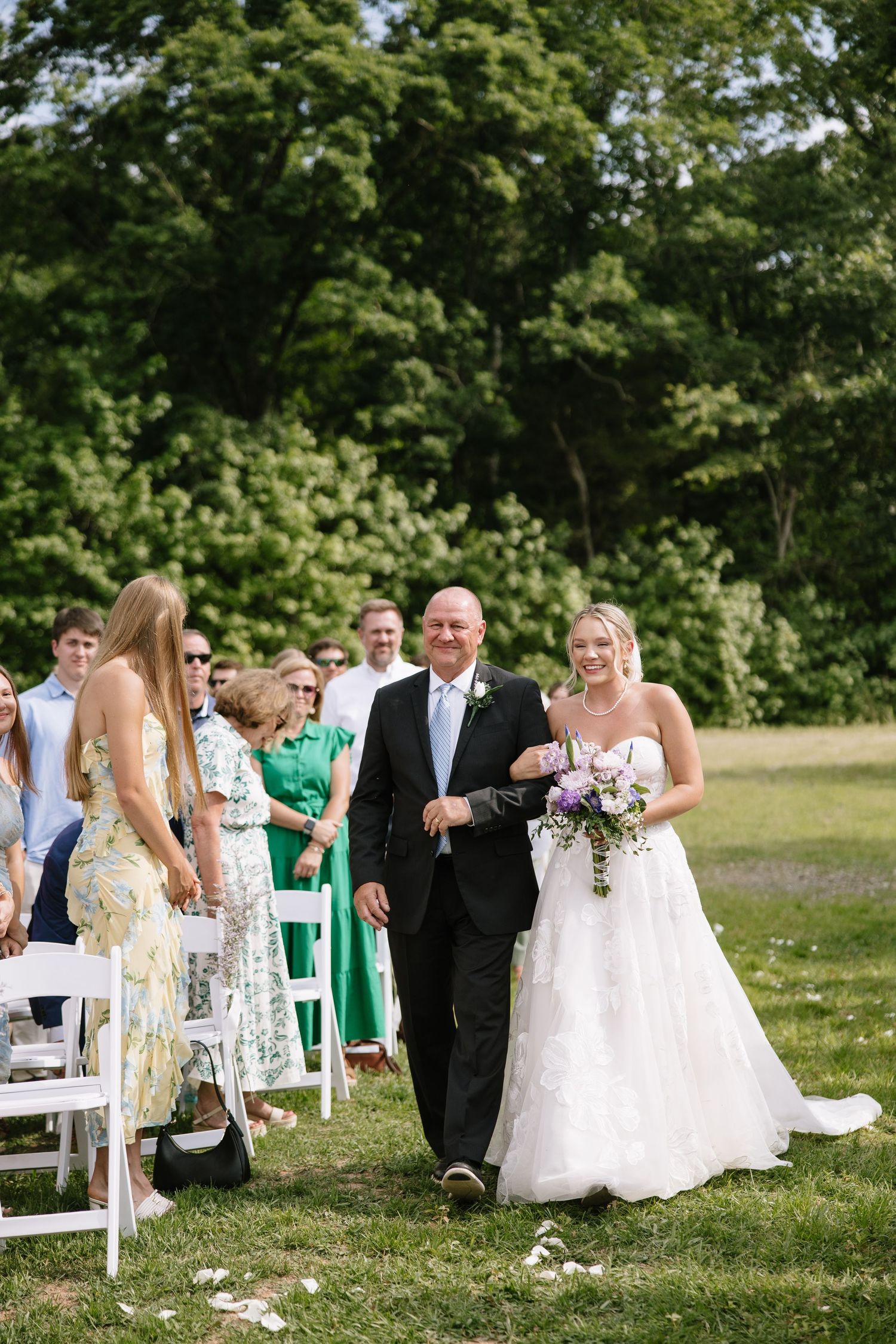 Bride walks down outdoor aisle with her father, holding purple bouquet at sunny wedding ceremony.
