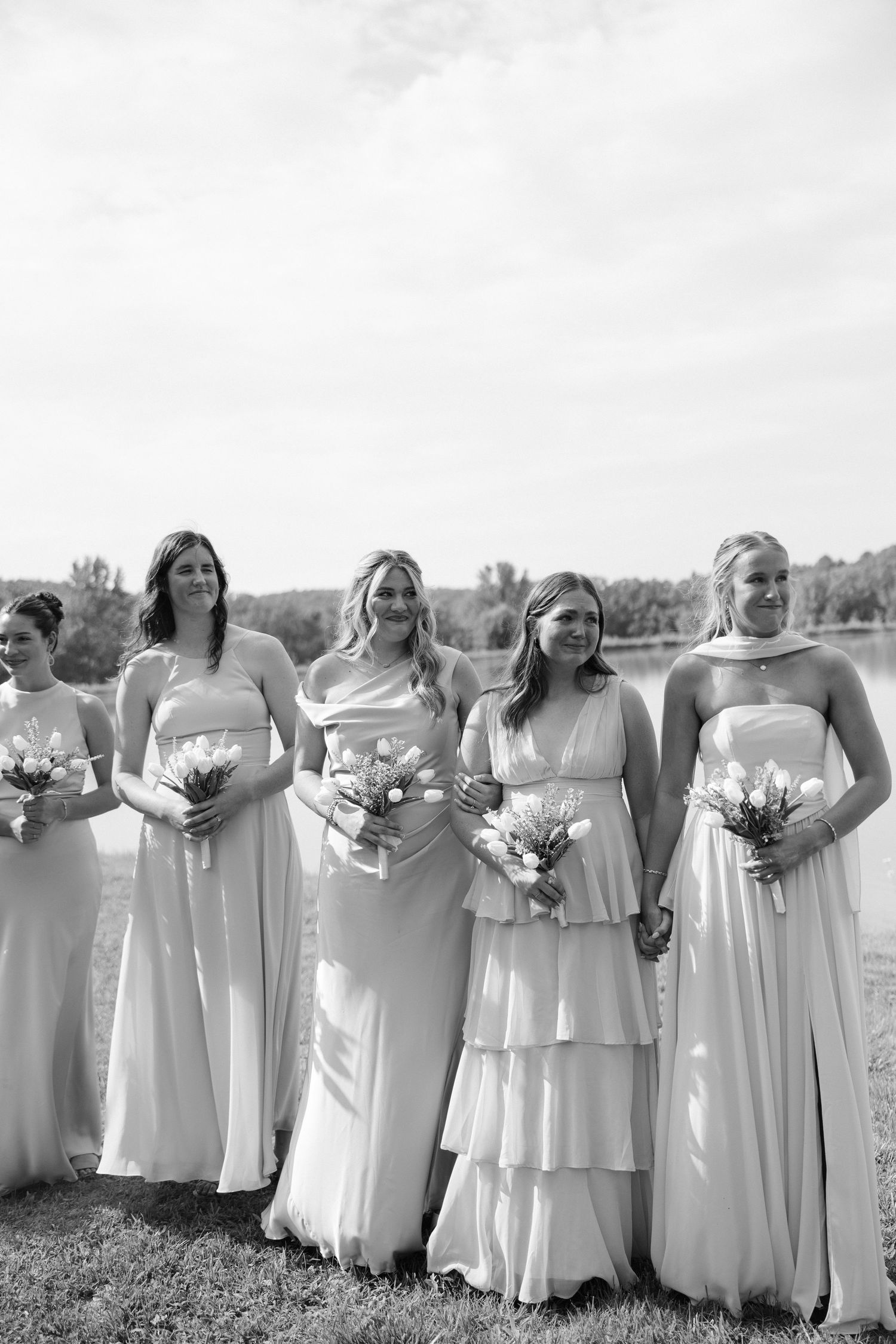 Five bridesmaids in long light-colored gowns holding floral bouquets pose outdoors in a black and white wedding photo.