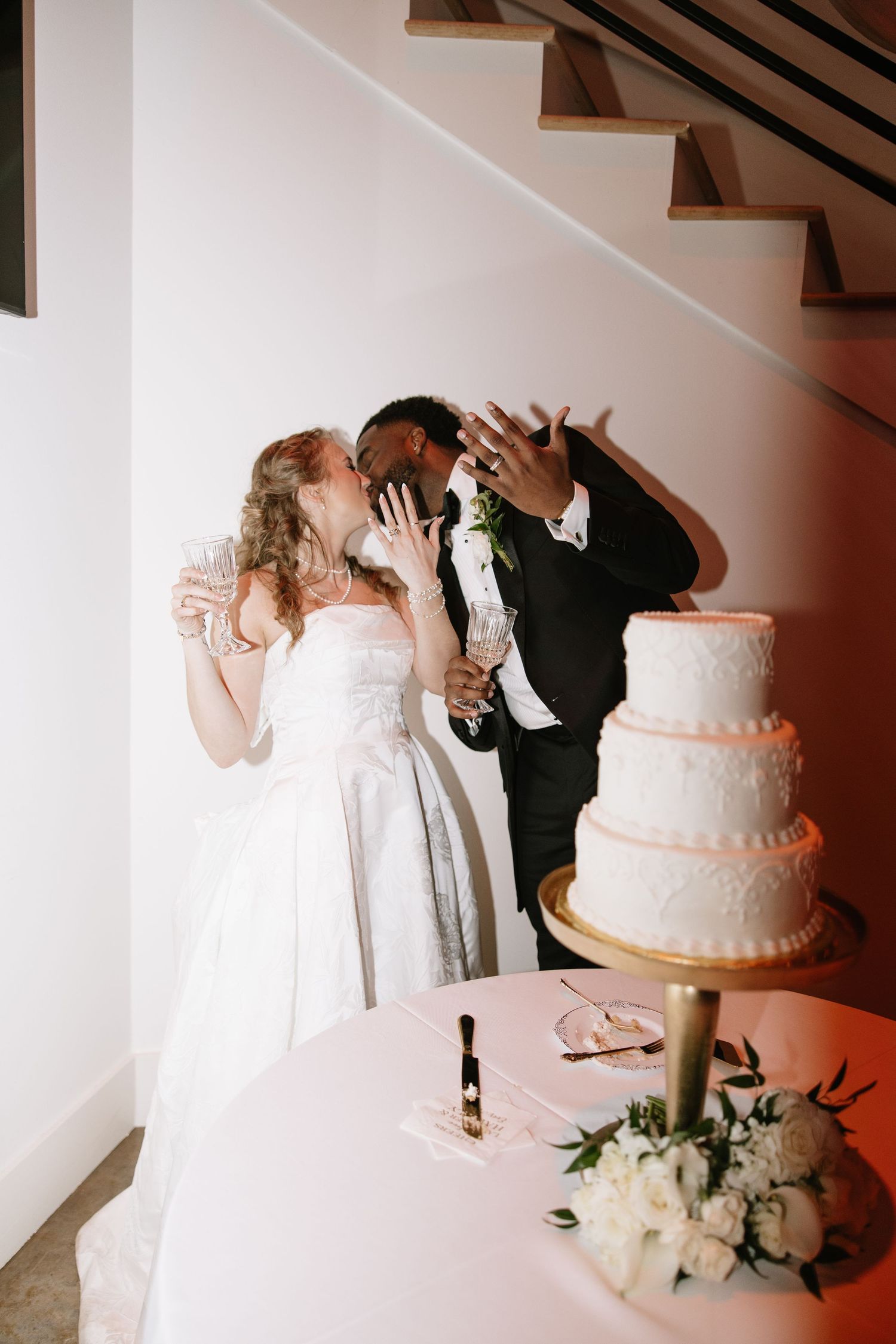 Newlyweds kissing and showing rings beside three-tier white wedding cake.