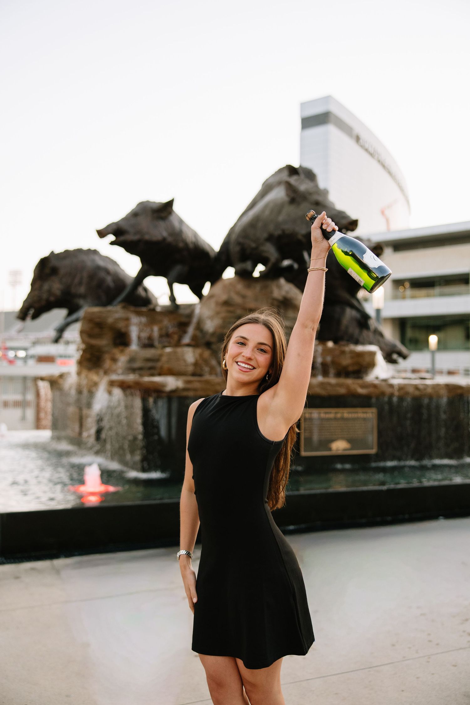 Celebrating graduate in black dress raising champagne bottle in front of Wild Band of Razorbacks fountain.