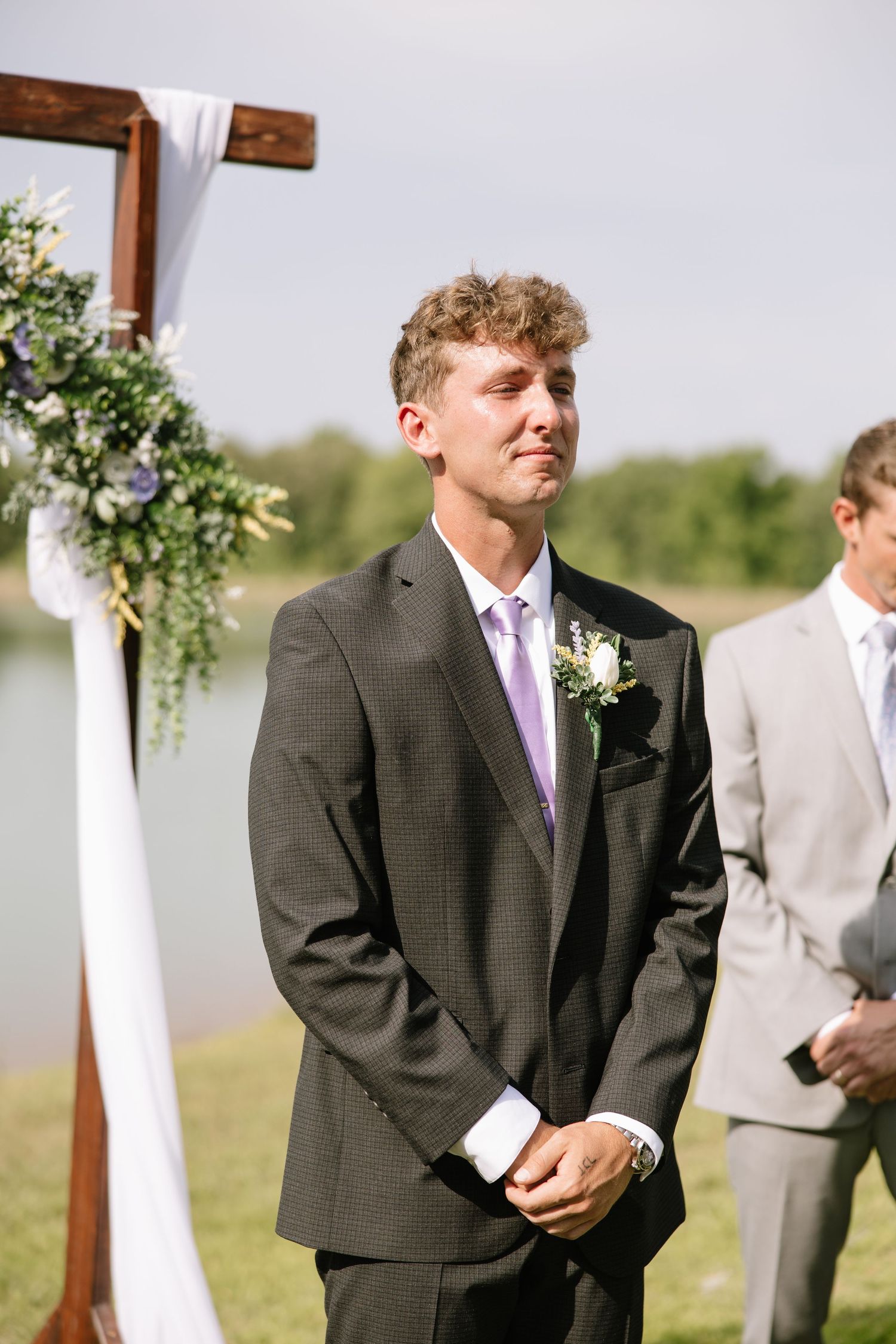 Emotional groom in dark suit with lavender tie reacting as bride walks down aisle at outdoor wedding.