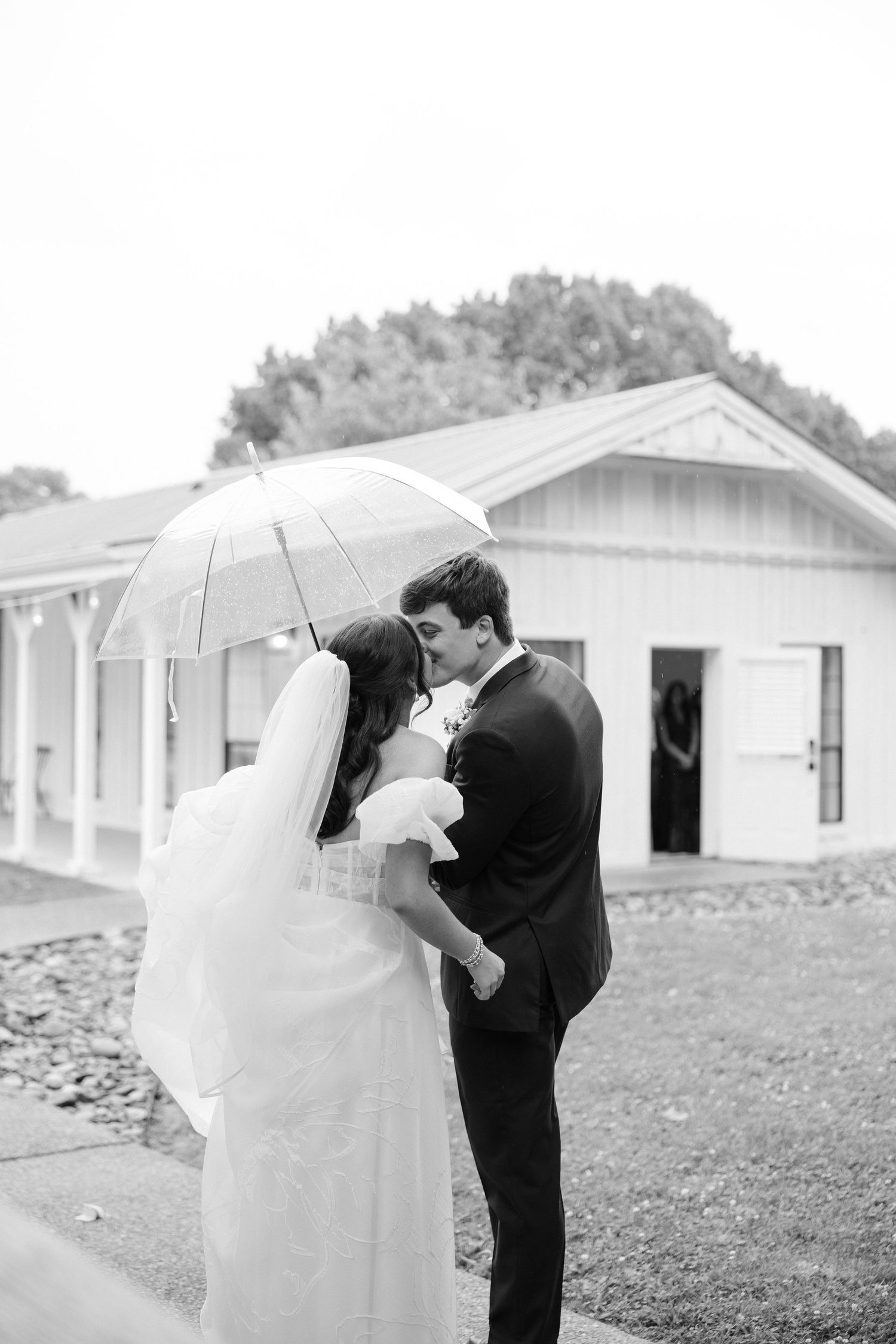 Black and white photo of a bride and groom sharing a kiss under a clear umbrella on their rainy wedding day.