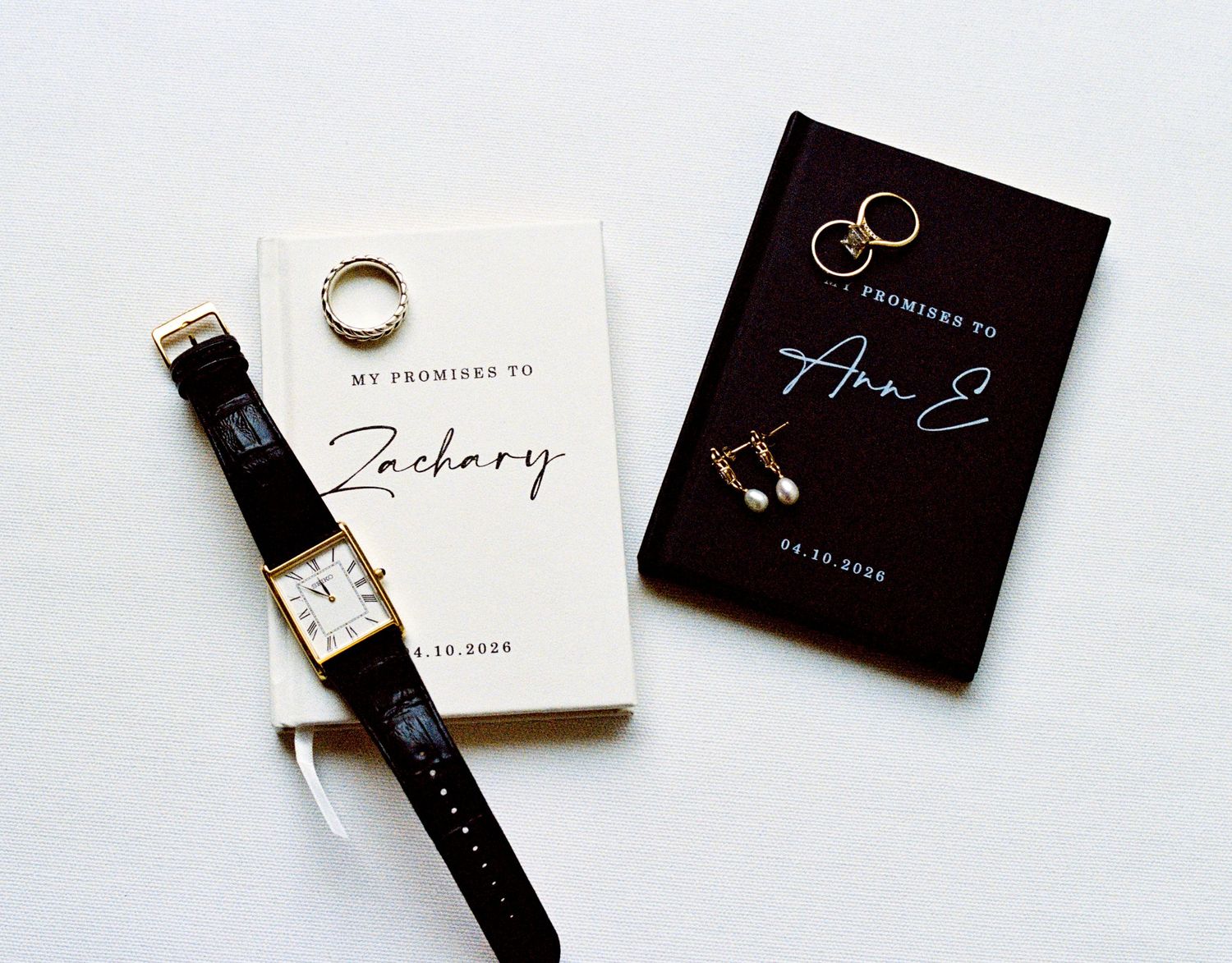 Wedding flat lay featuring vow books, gold watch, pearl earrings, and ring on white background.