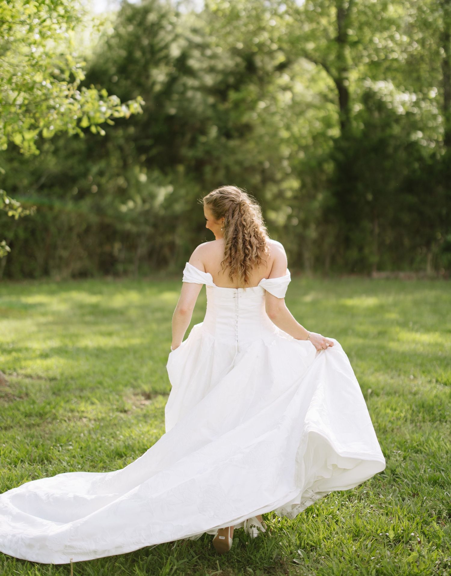 Bride in off-shoulder white wedding gown walking through lush green garden.
